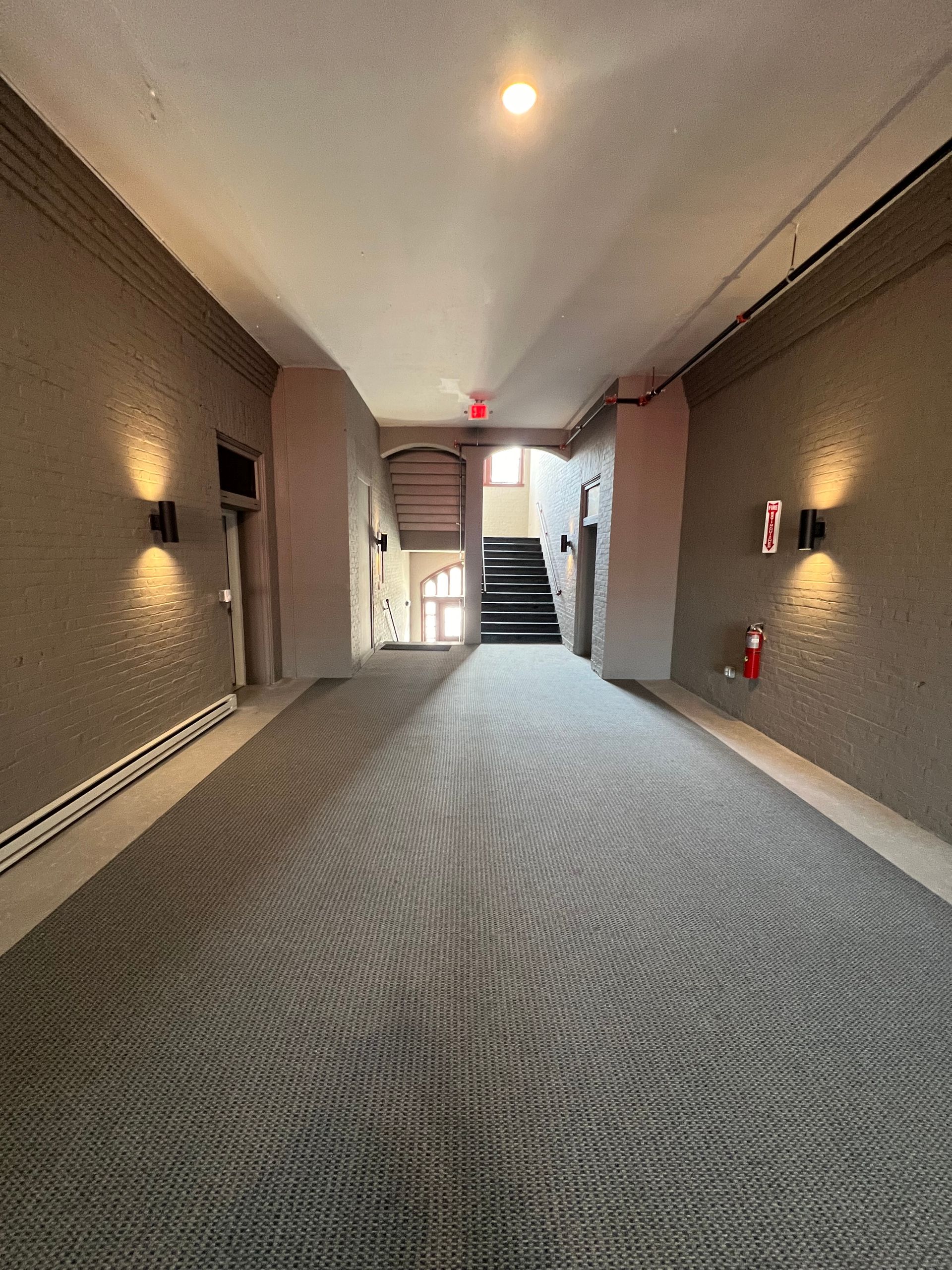 Hallway with gray textured walls and patterned carpet, leading to stairs. Two sconces and a fire extinguisher.