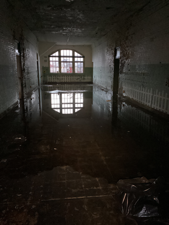 Flooded hallway in an abandoned building. Water reflects a barred window at the end. Dark, moldy interior.