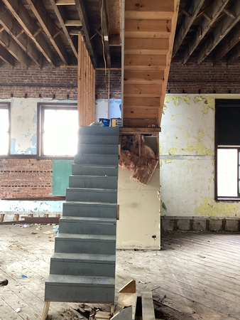Interior view of a building under renovation, showing stairs, exposed ceiling beams, and damaged walls.