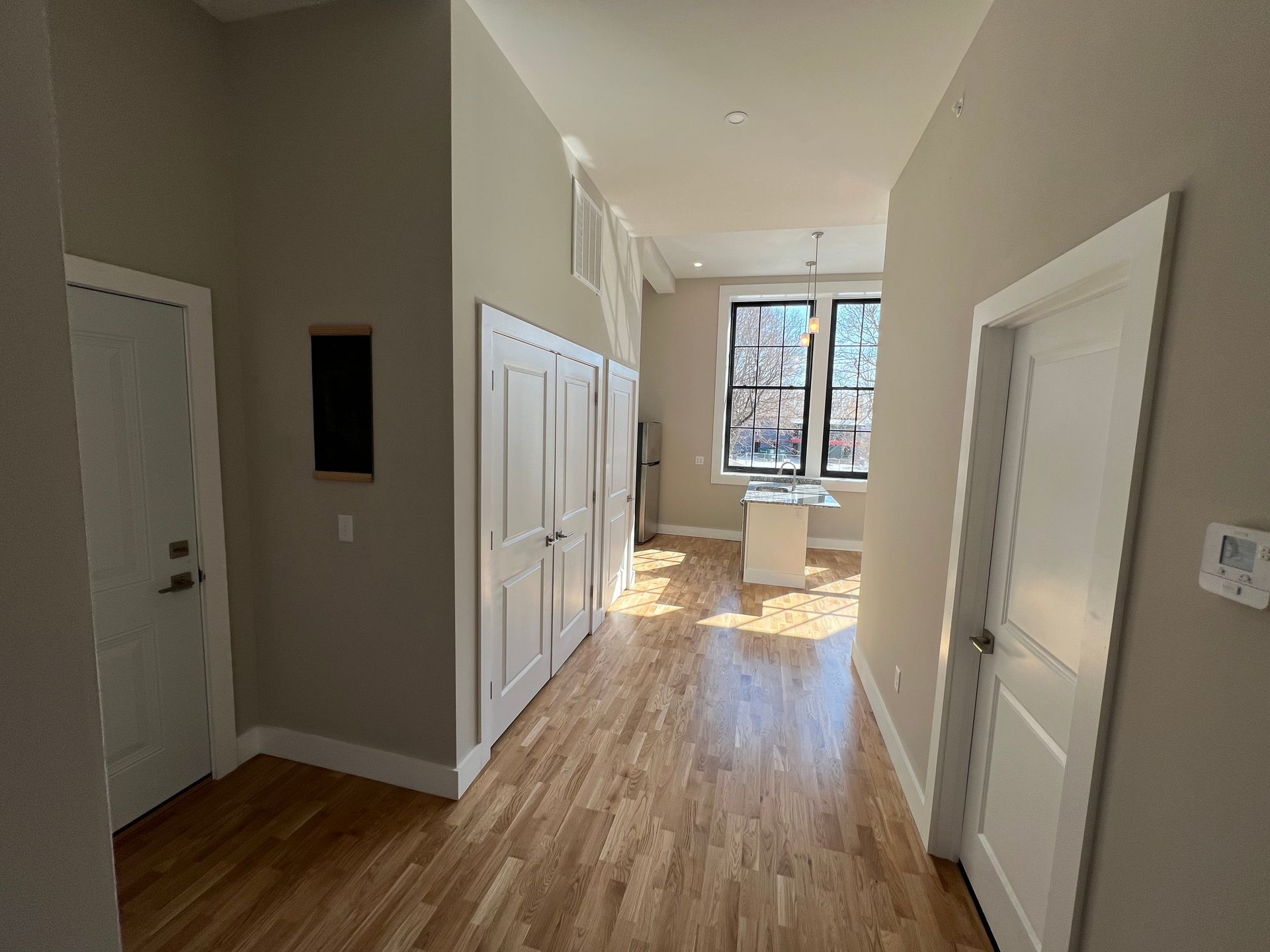 Hallway with hardwood floor, doors on both sides, leading to a kitchen with a large window.