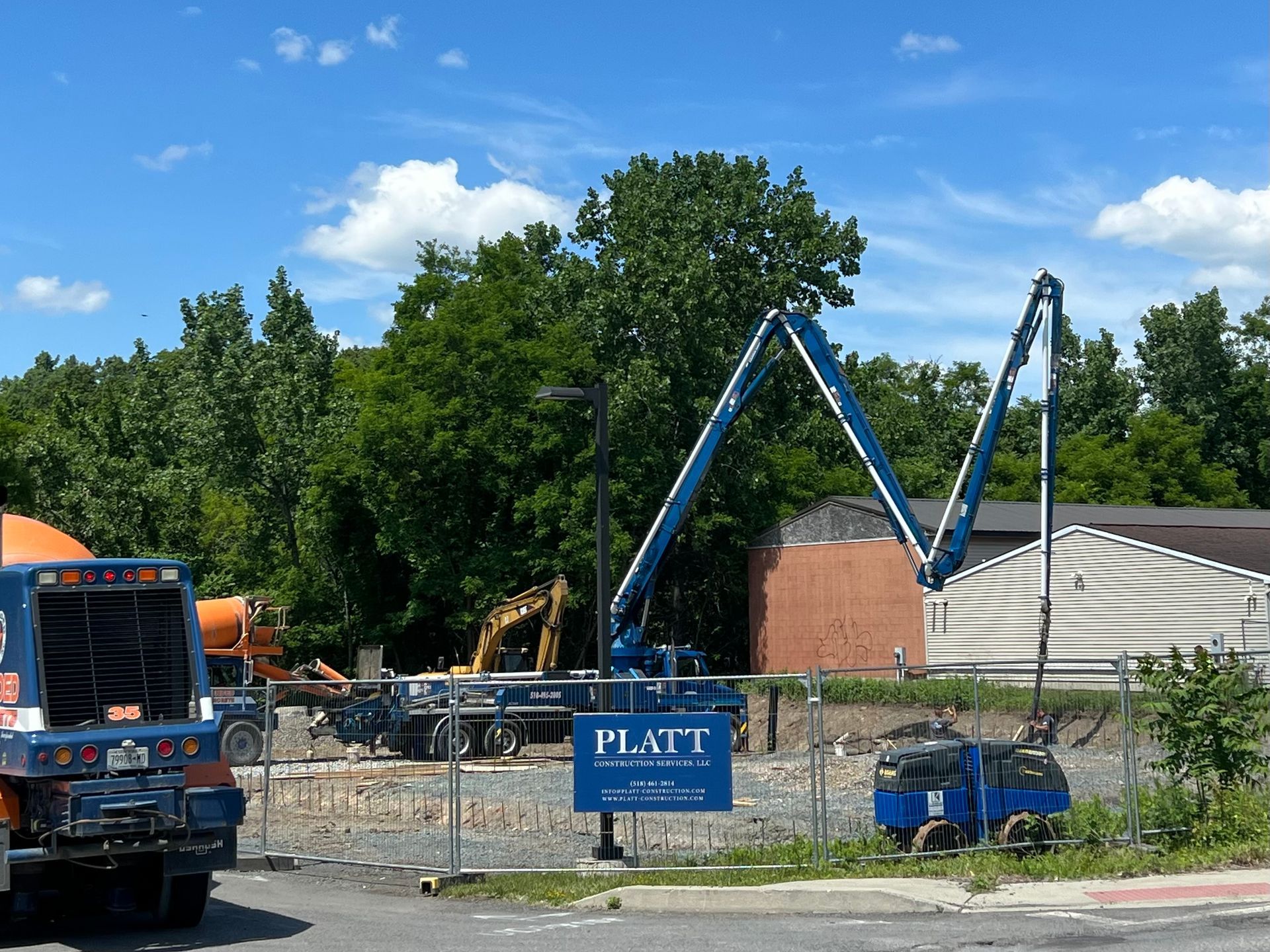 Construction site with concrete pump, mixer truck, and excavator. Trees in the background, blue sky.
