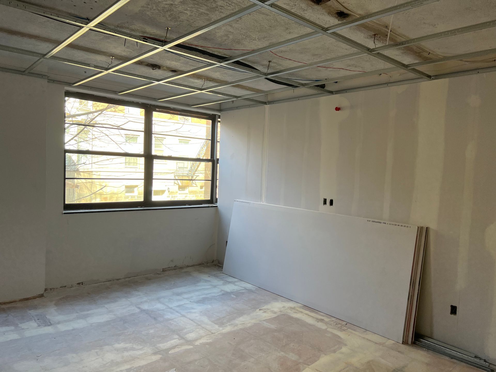 Unfinished room with drywall walls and ceiling, window, exposed metal grid, and sheetrock on the floor.