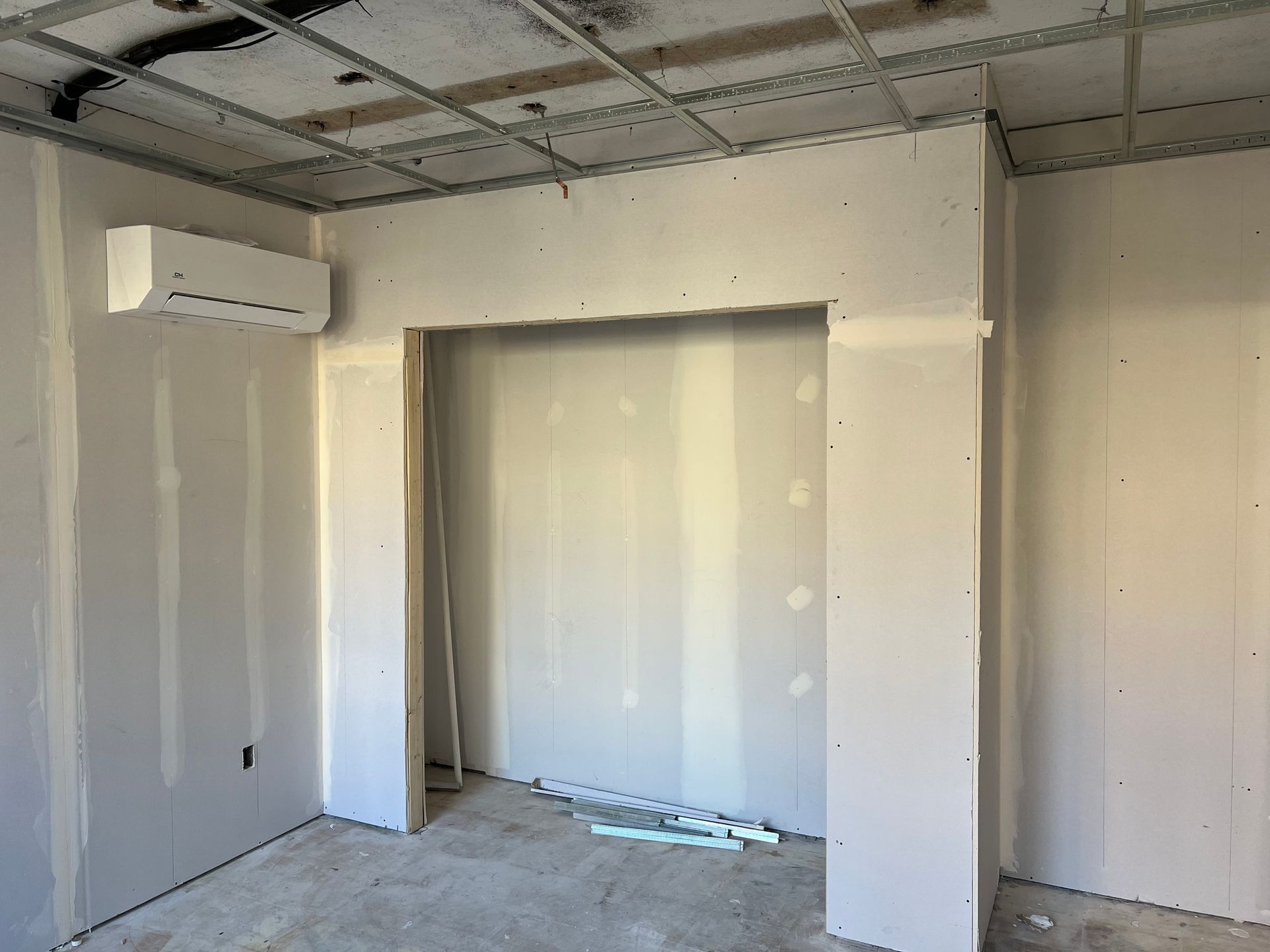Interior room under construction, drywall on walls, exposed ceiling grid, air conditioning unit, doorway.
