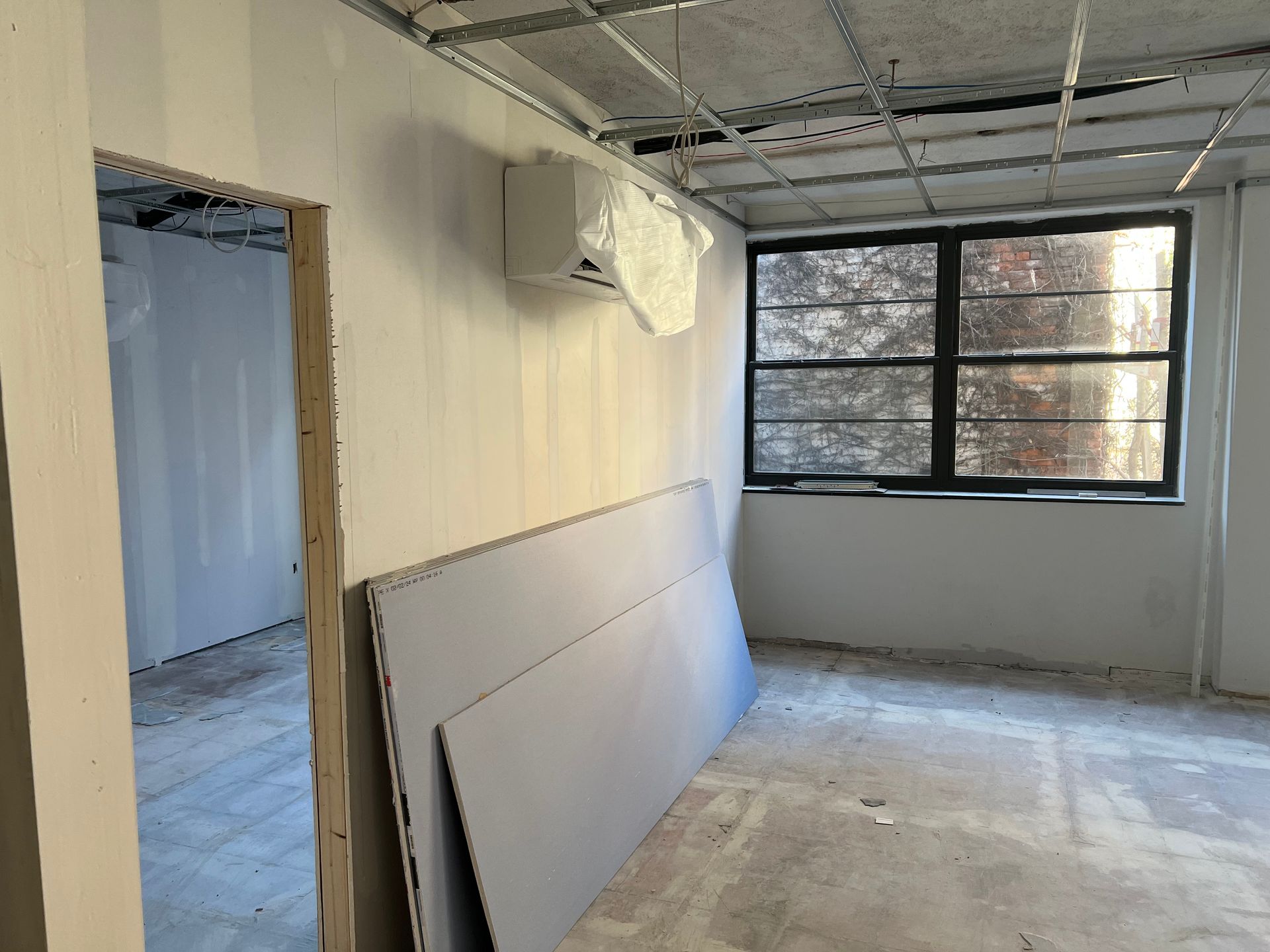 Interior of a room under construction, with drywall leaning against a wall, an open doorway, and a window.
