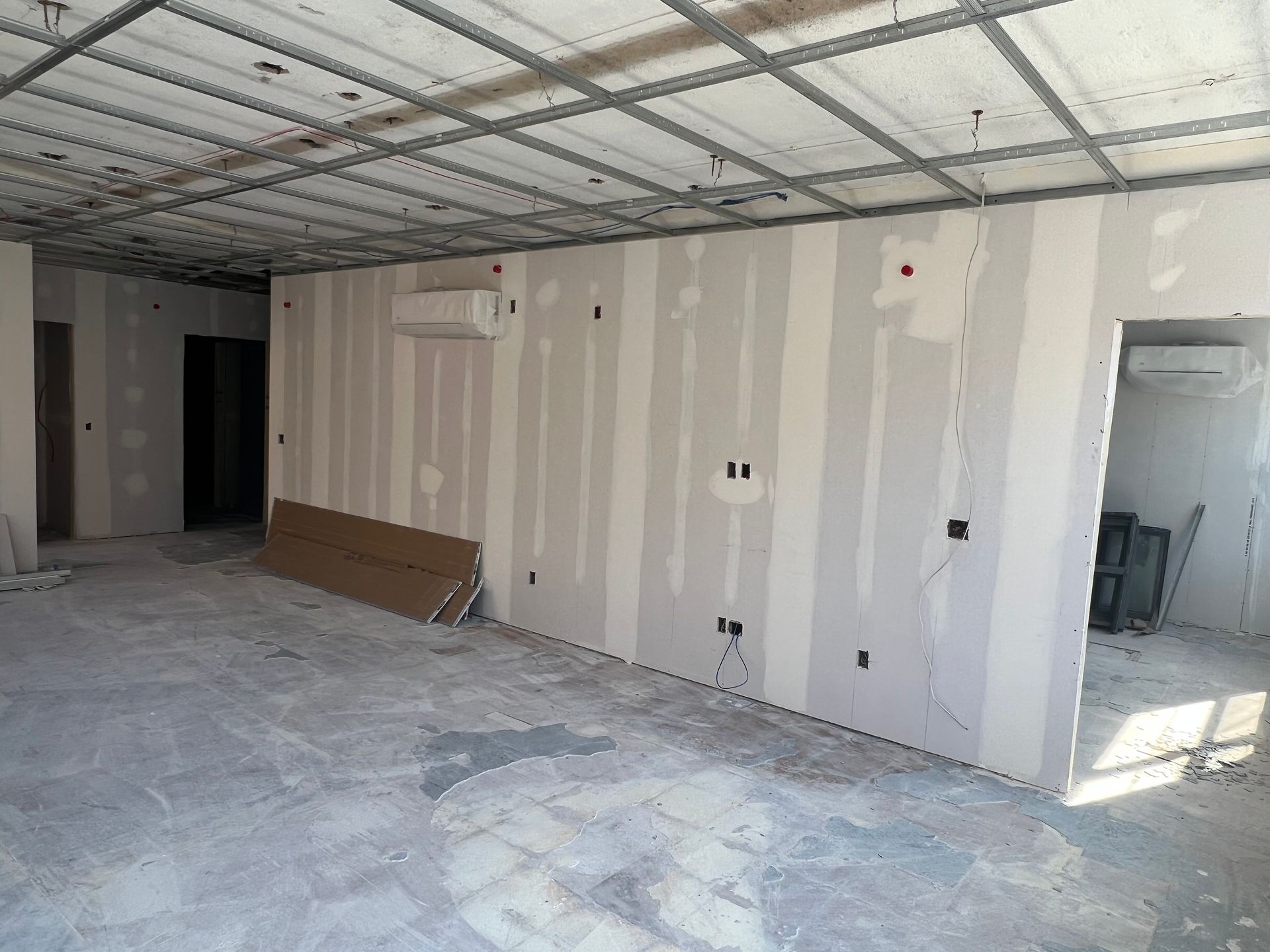 Interior under construction: drywall on walls, exposed ceiling grid, concrete floor.
