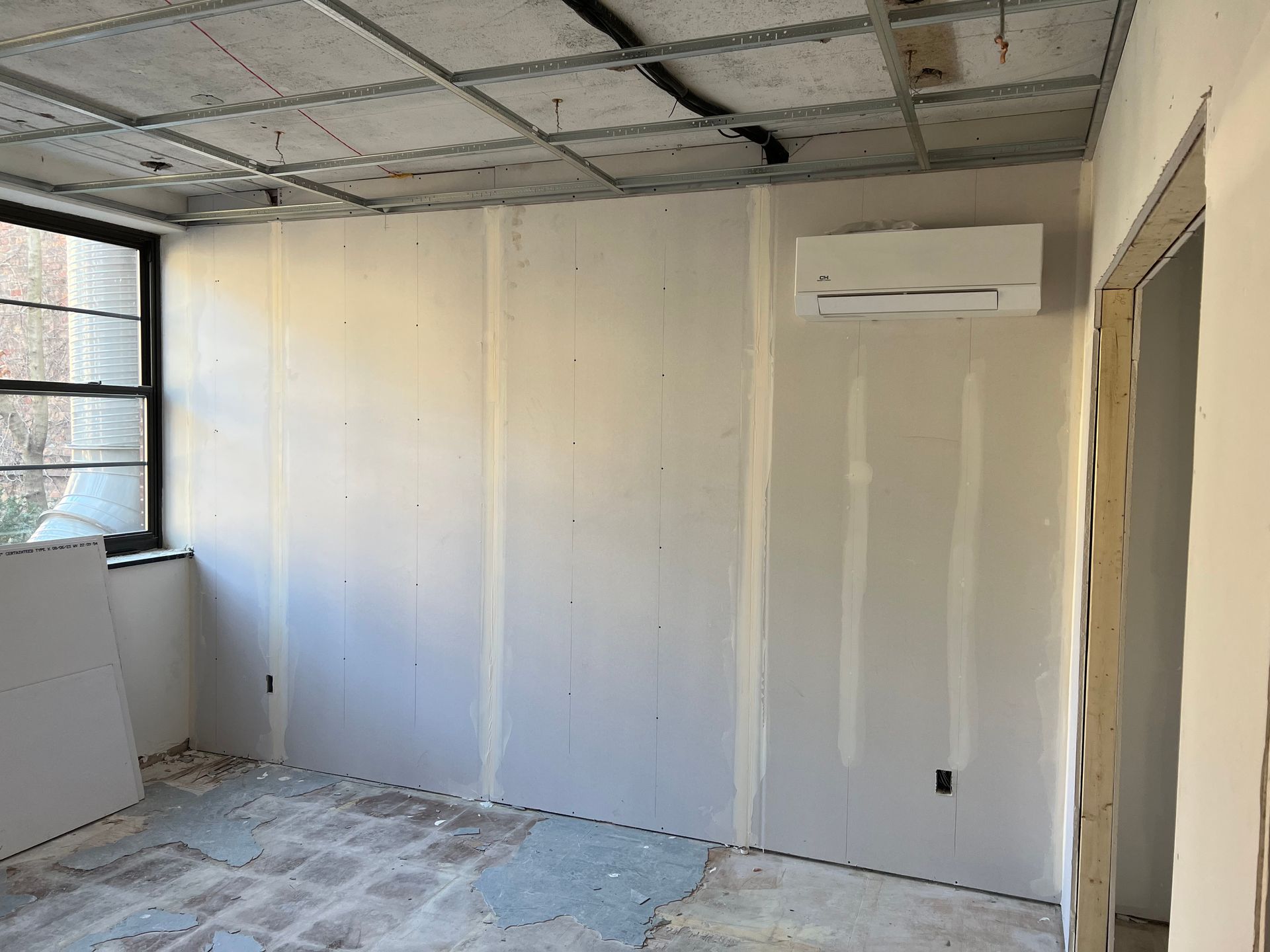 Room under construction: drywall on walls, air conditioning unit, unfinished ceiling, window, and doorway.