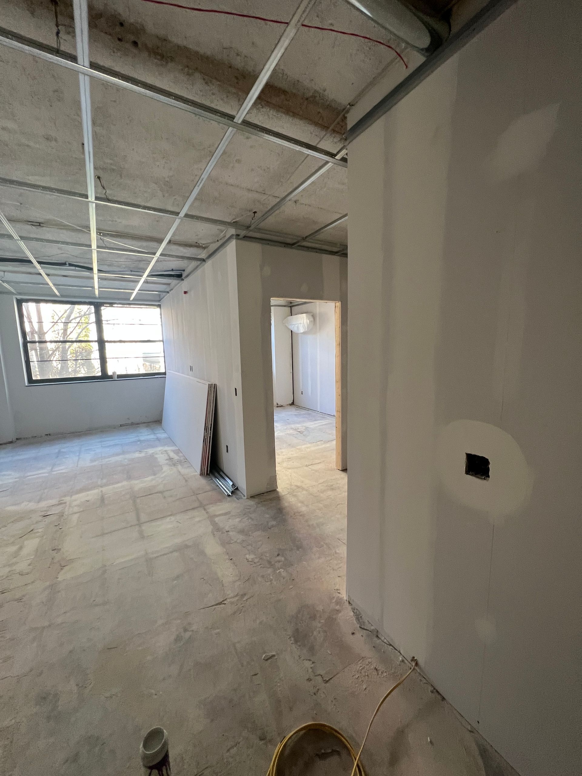 Interior of a room under construction; bare concrete floors and walls, metal ceiling grid, doorway to another room.