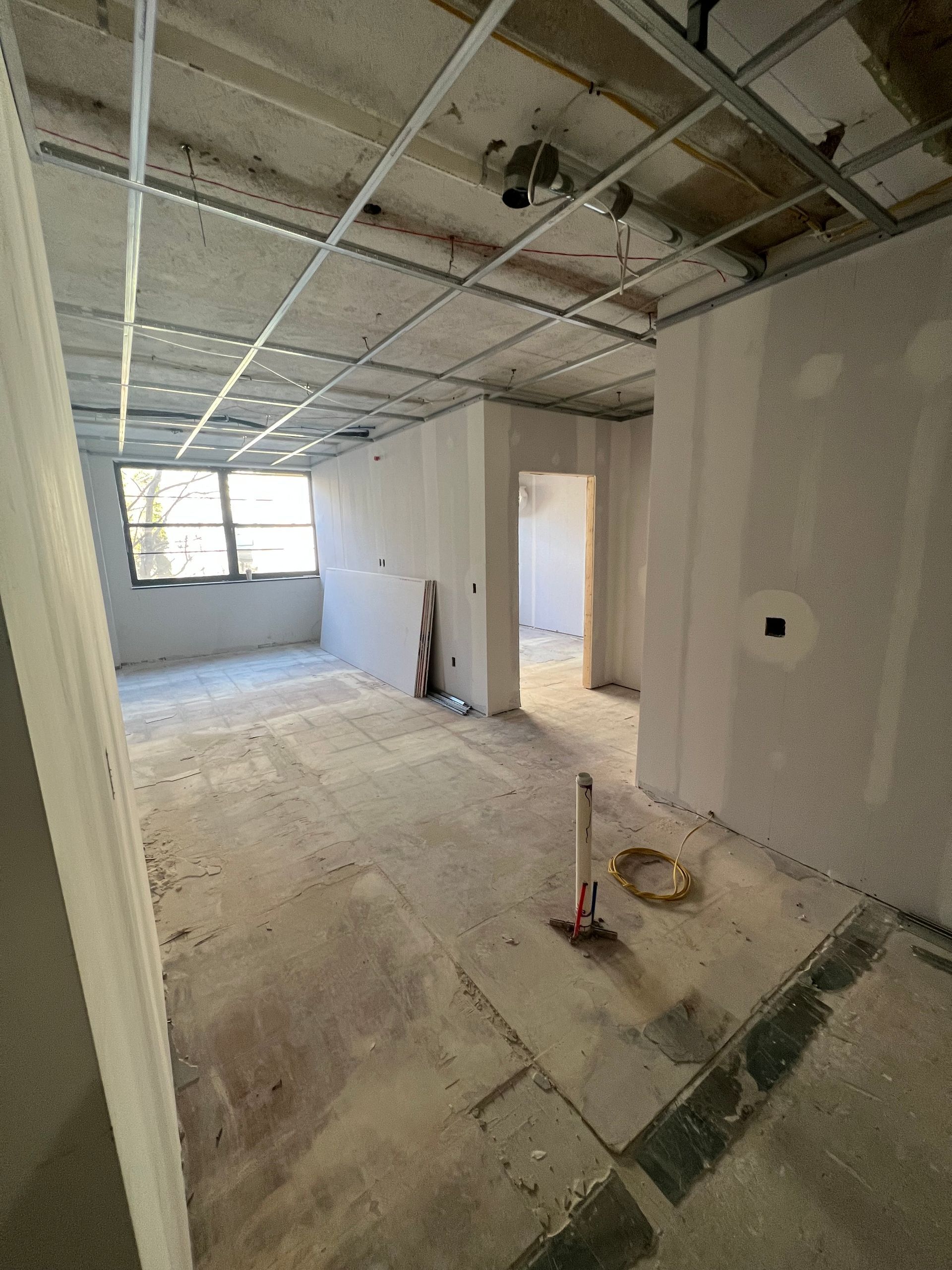 Interior room under construction with drywall walls, exposed ceiling grid, concrete floor.