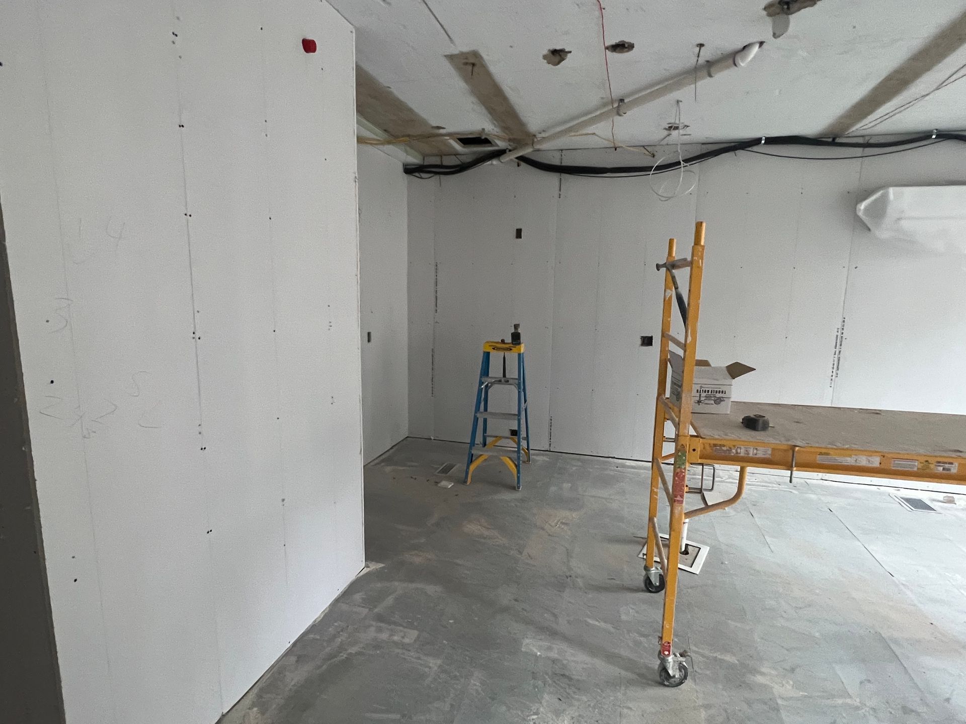 Interior construction site. Drywall walls and ceiling, scaffolding, and a ladder are present.