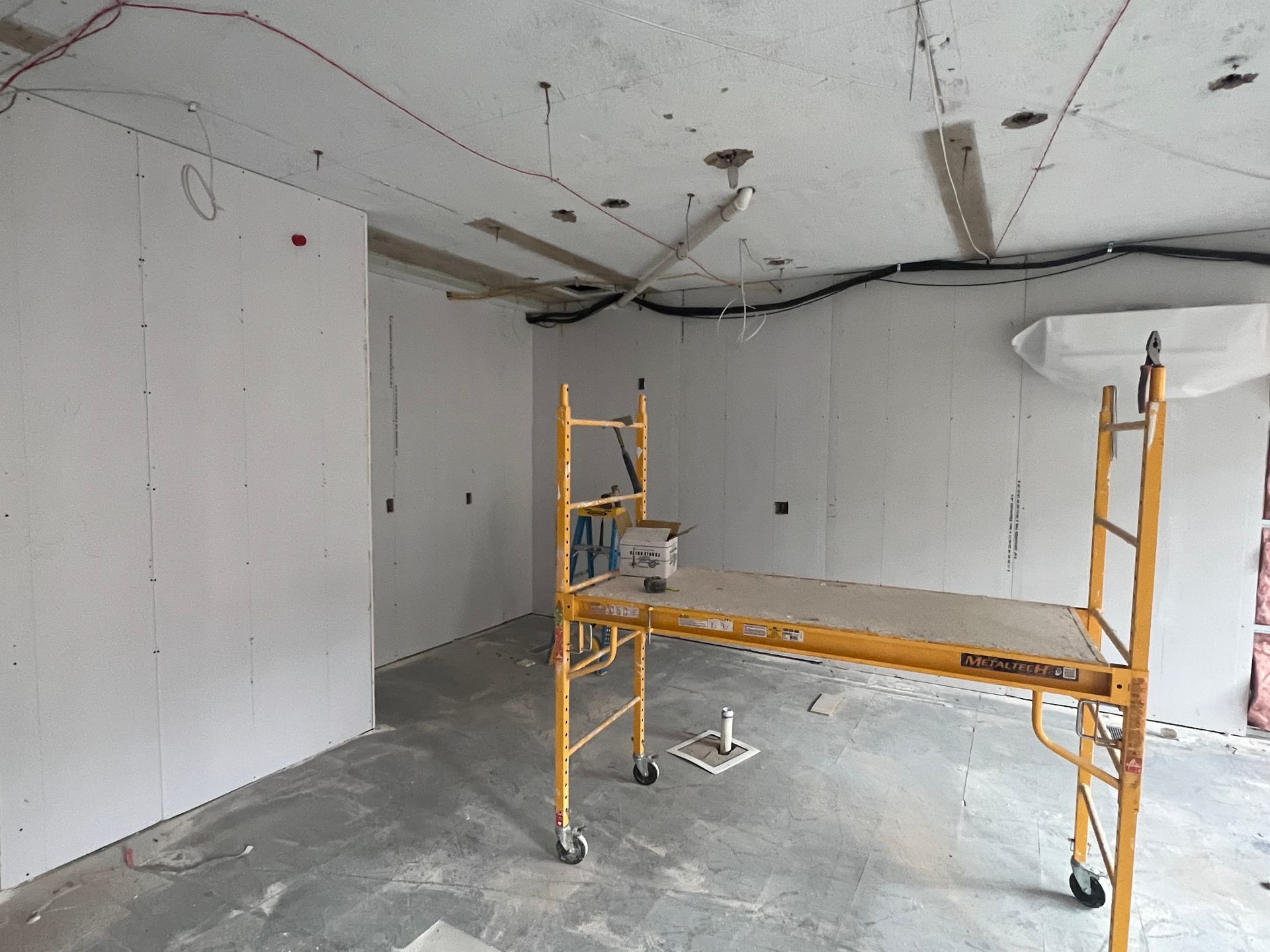 Interior of a room under construction with drywall and scaffolding, showing electrical wiring and a concrete ceiling.