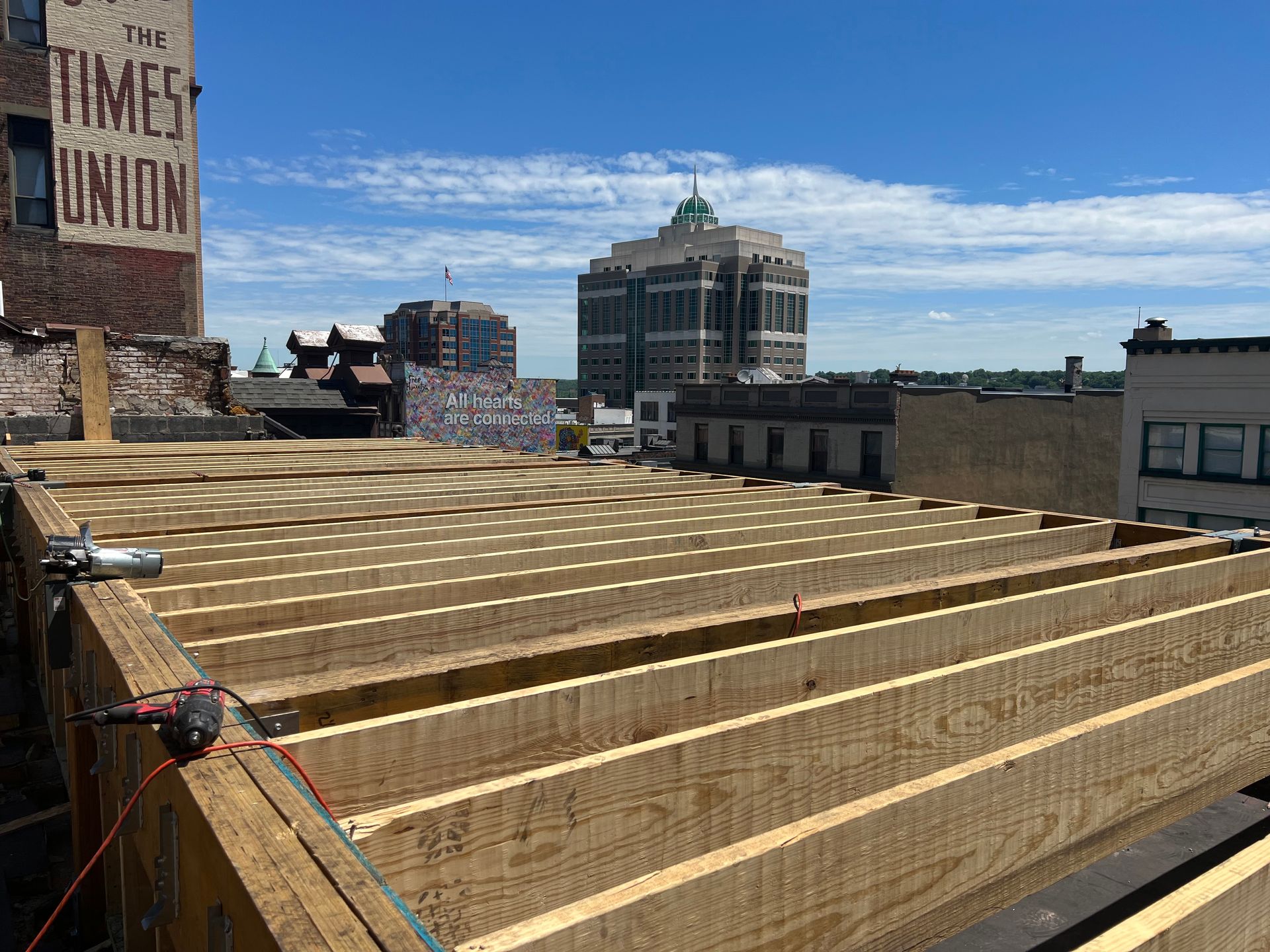 Rooftop view of wooden beams being installed, with city buildings in the background under a blue sky.