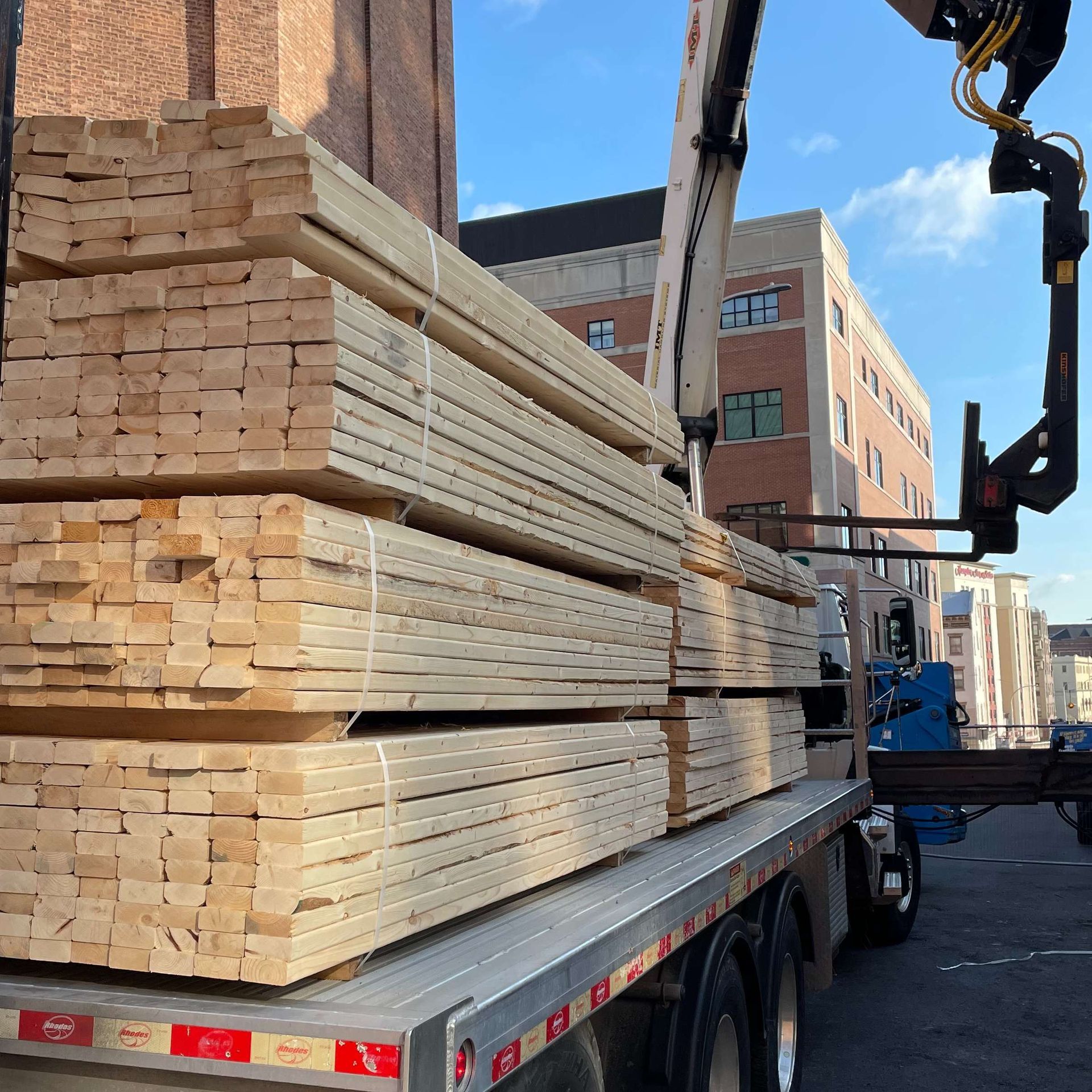 A truck bed loaded with lumber being lifted by a crane in front of a city building.
