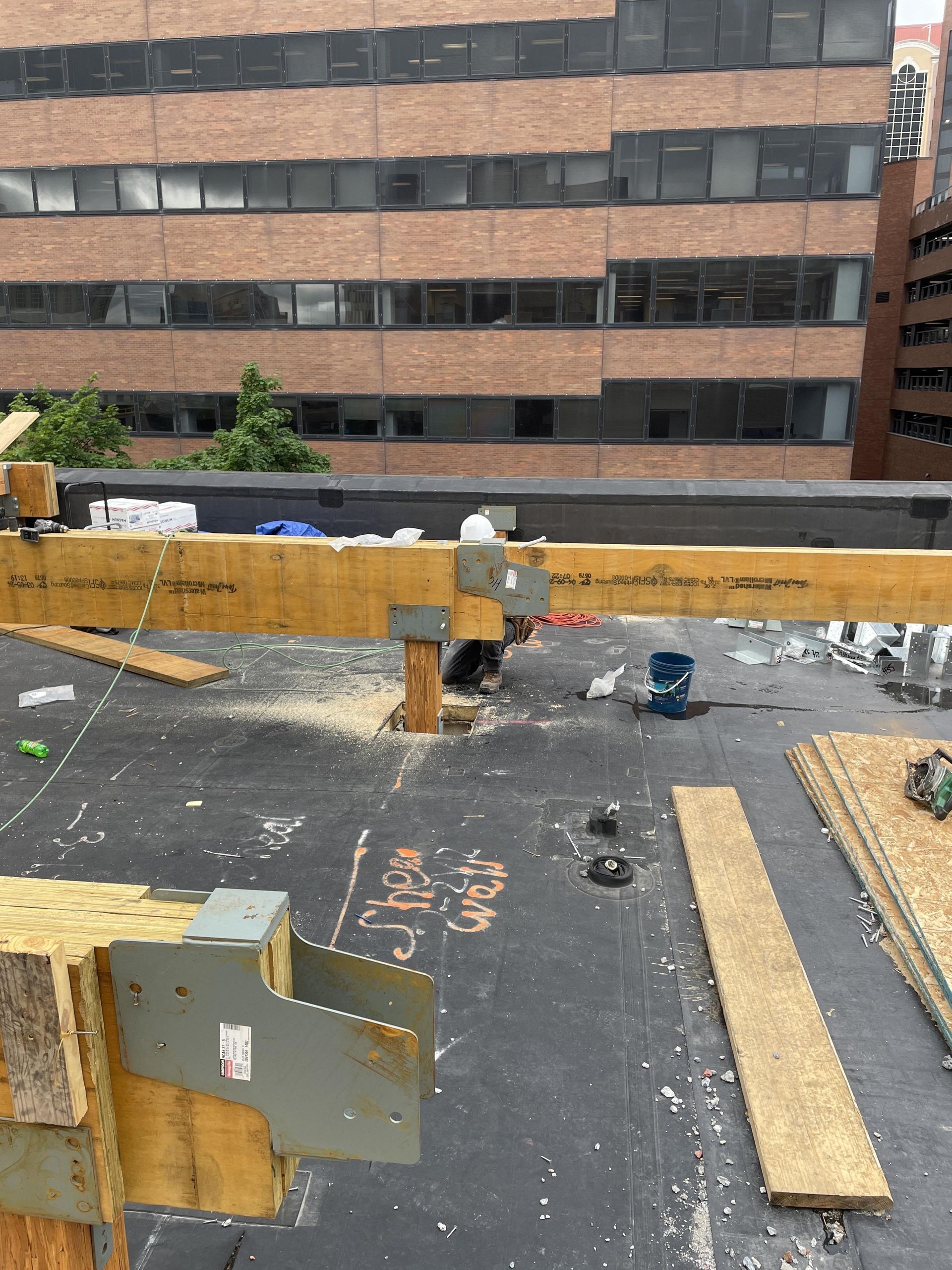 Rooftop construction with wooden beams, metal brackets, and flat roof. Building in the background.