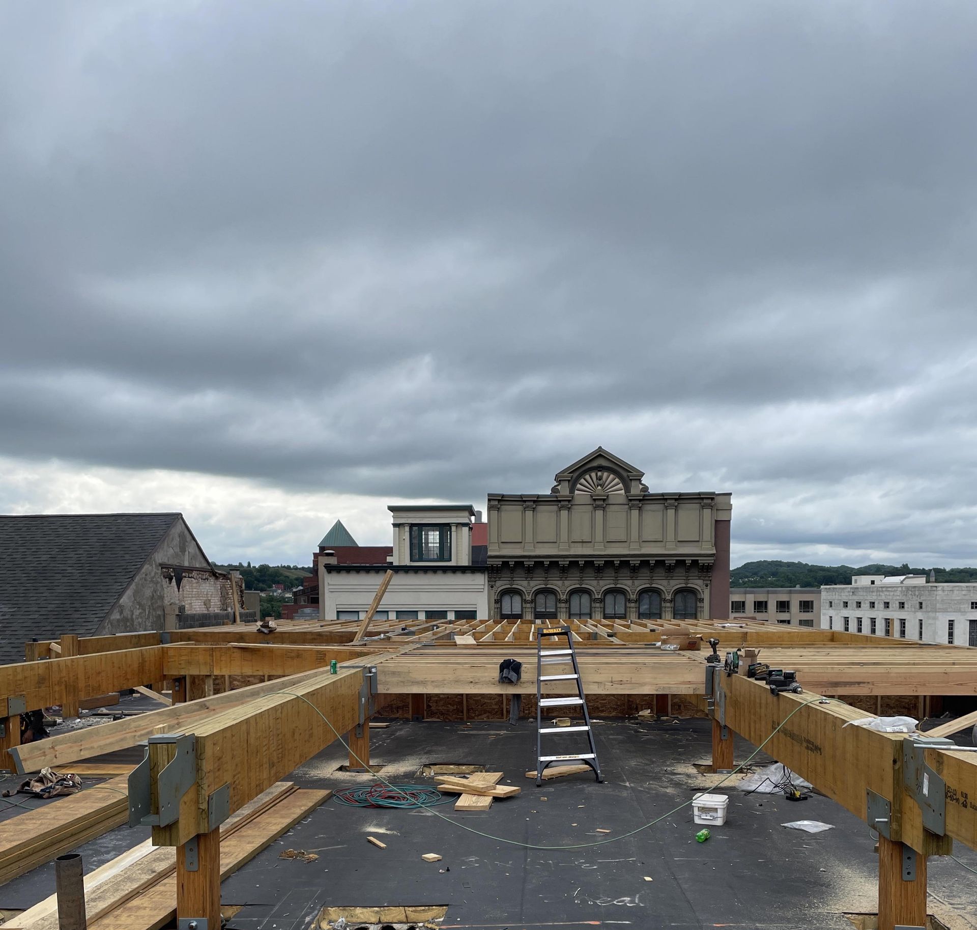 Construction site on a building's roof with wooden beams, ladder, and cloudy sky. Other buildings in background.