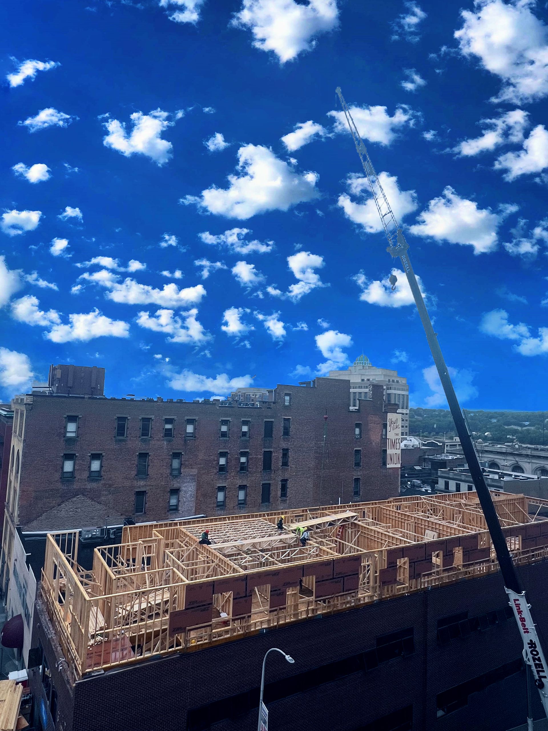 Construction site with crane, wood framing, and a brick building against a blue sky with clouds.