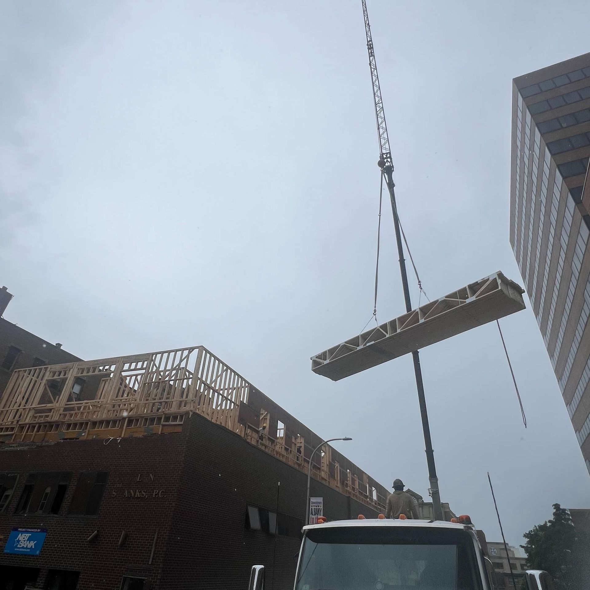 Crane lifting a wooden structure above a building under construction on a city street. Cloudy sky.
