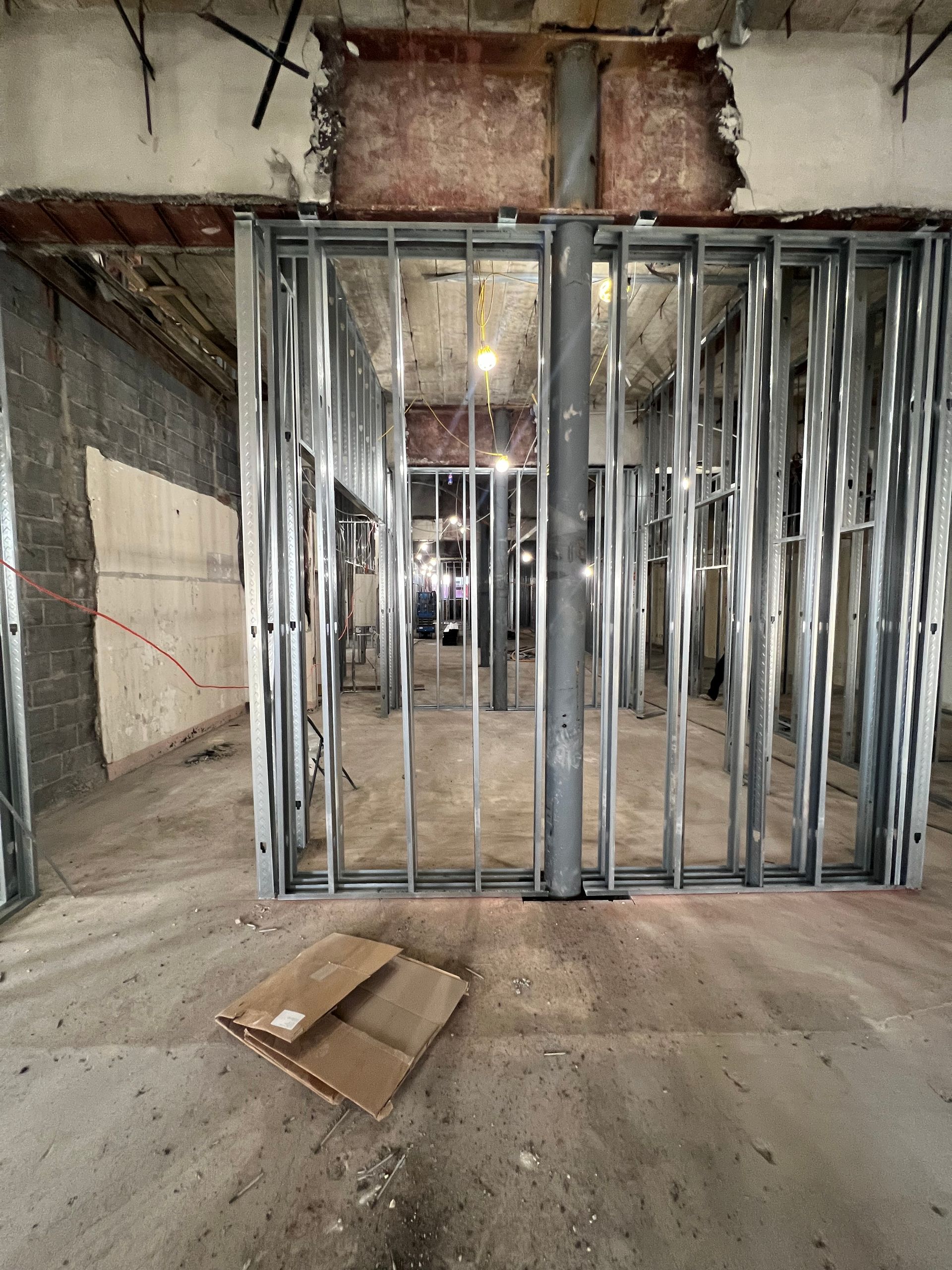 Interior construction site. Metal studs frame walls, with exposed pipes and damaged concrete overhead. Cardboard box on the floor.