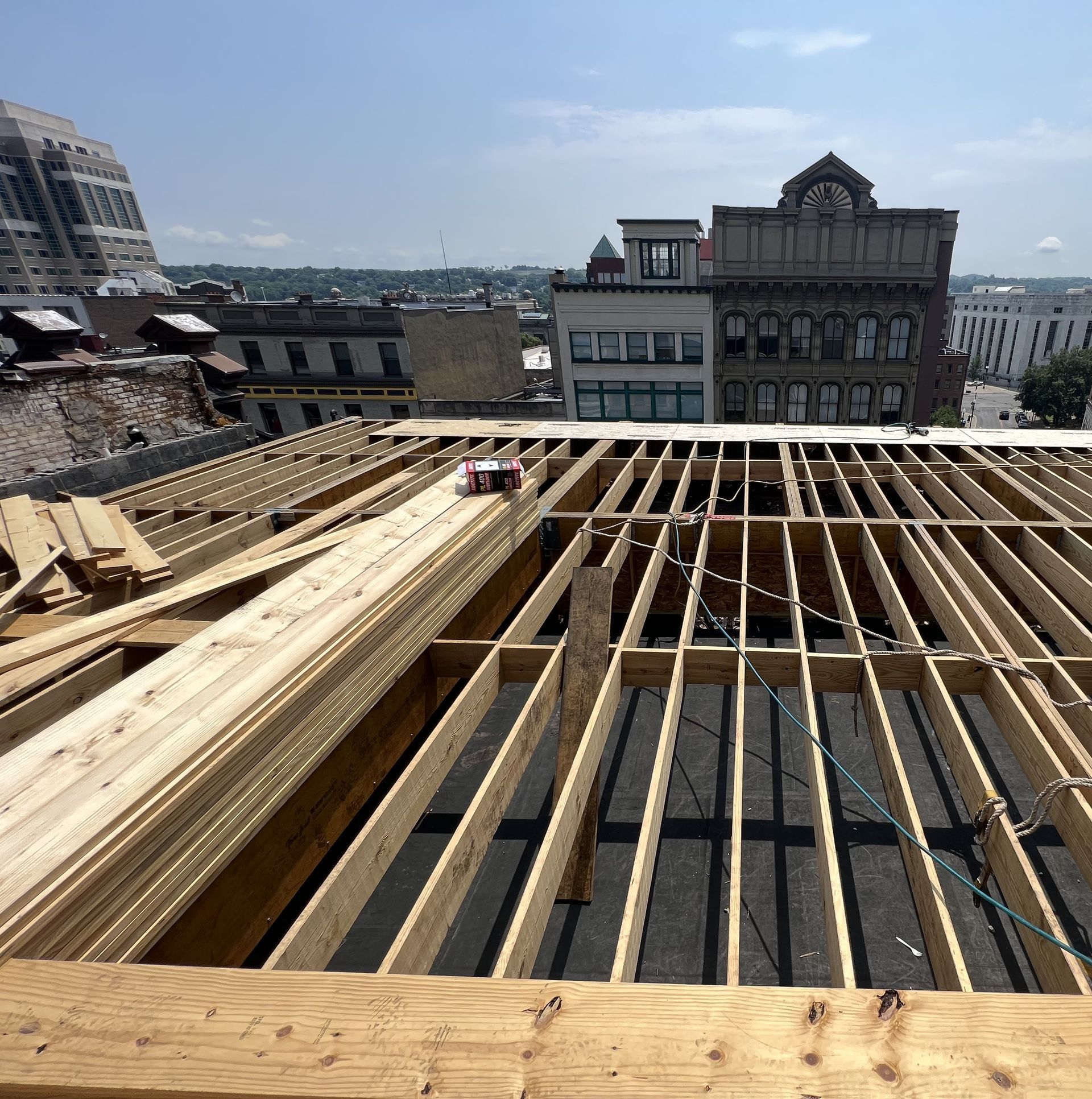 Rooftop view of wooden beams being laid for a roof construction project, city buildings in the background.