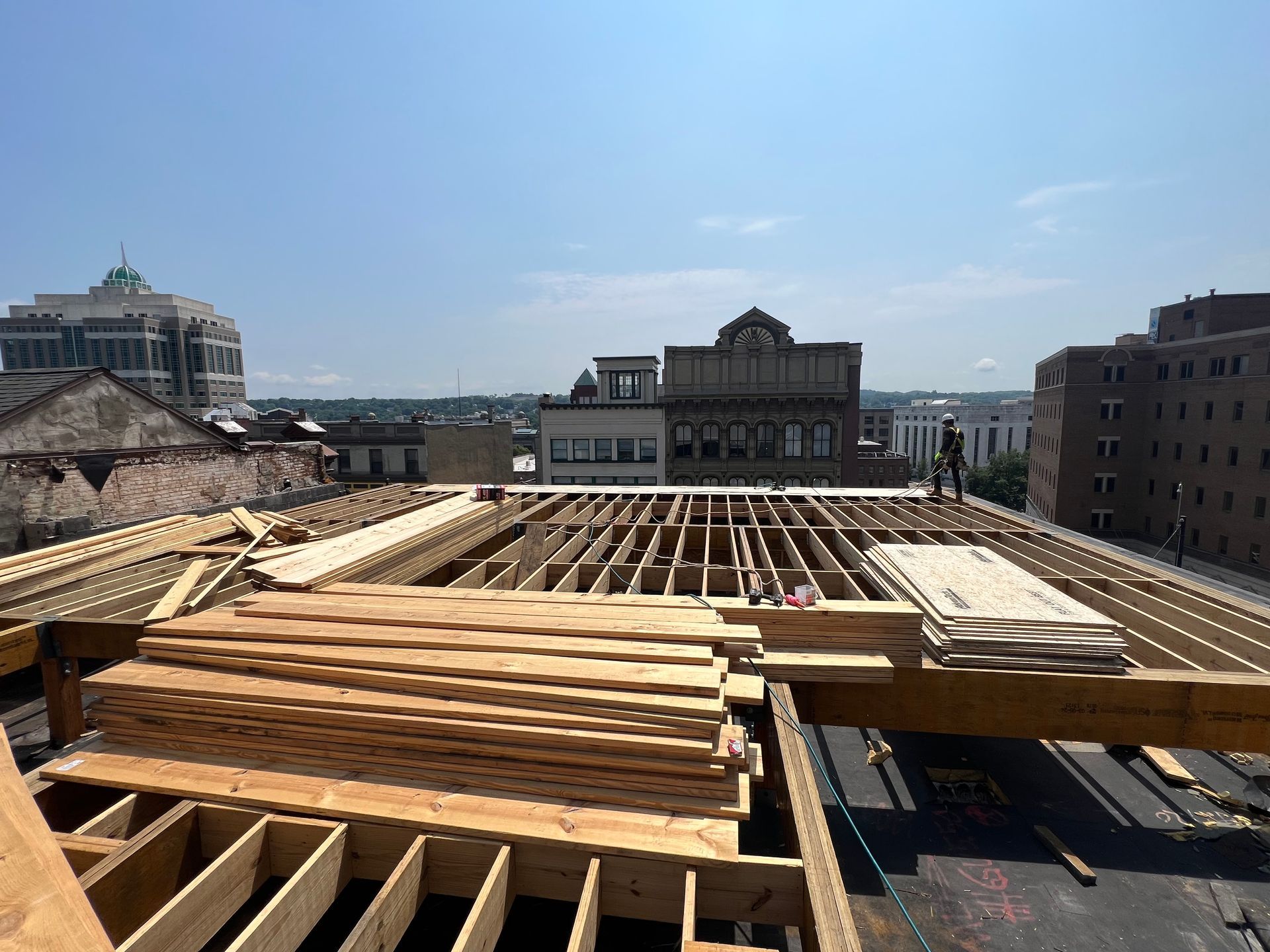 Rooftop construction with exposed wooden beams and stacked lumber; city buildings in the background under a blue sky.