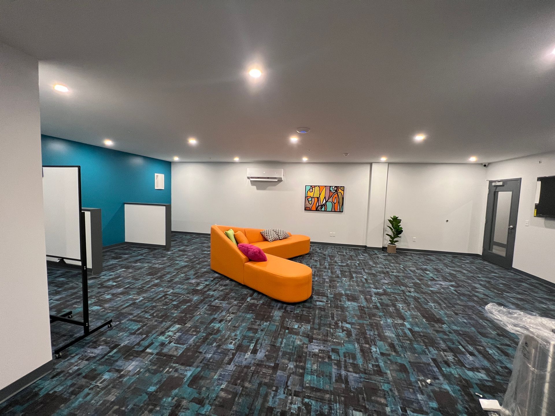 Open-plan room with an orange sofa, colorful artwork, and a teal wall. Blue and gray carpet.