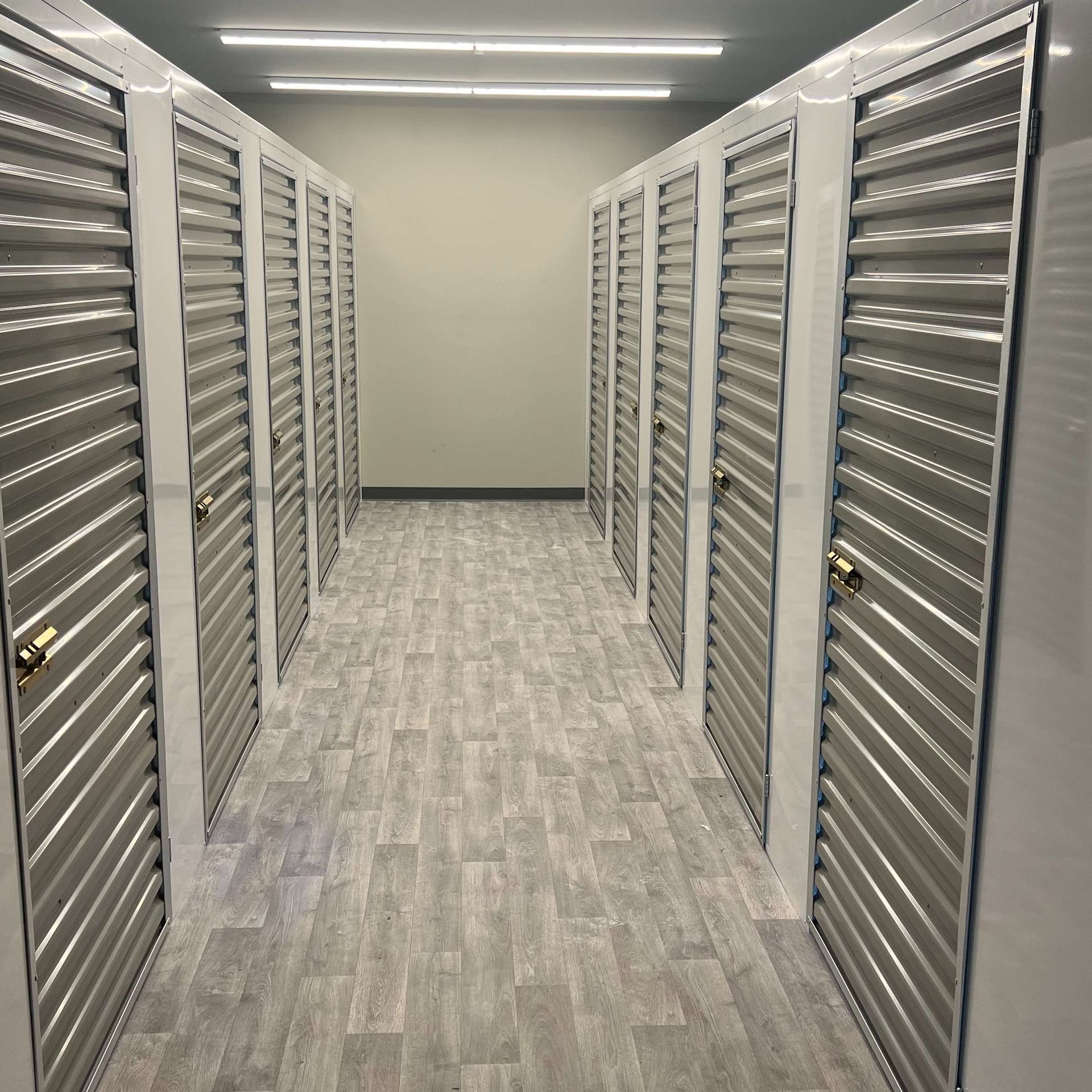 Storage unit hallway with rows of metal doors and a wood-look floor.