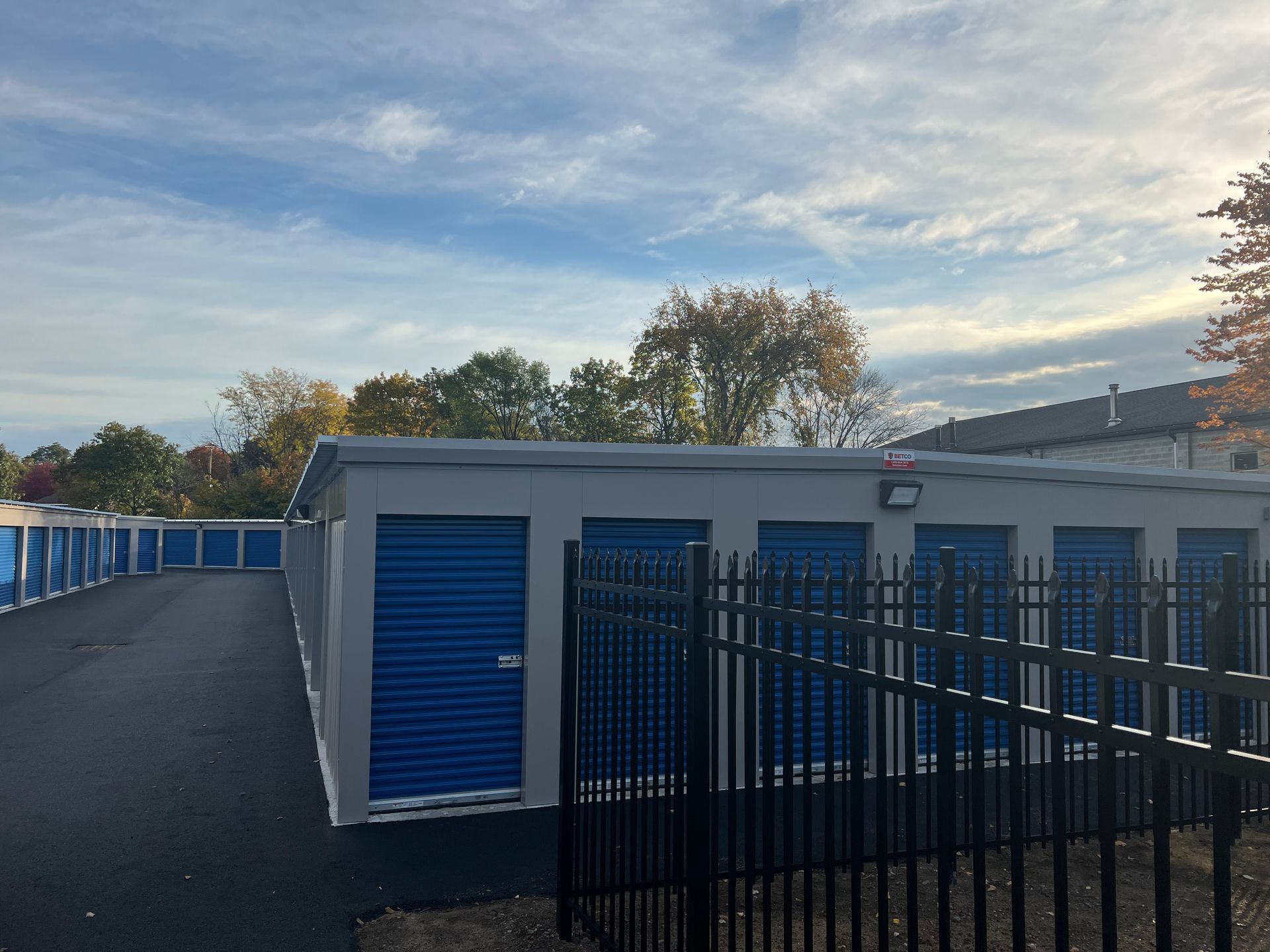 Exterior view of a storage facility with gray walls, blue doors, and a black fence under a cloudy sky.
