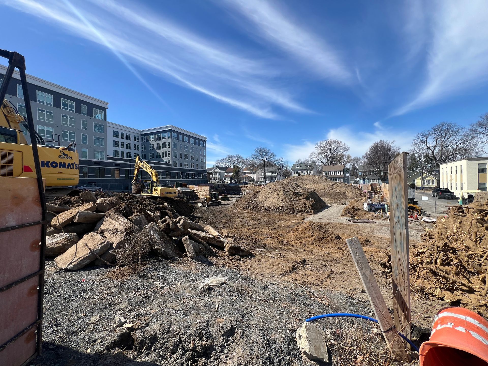 Construction site with excavators, debris, dirt piles, and buildings under a blue sky.