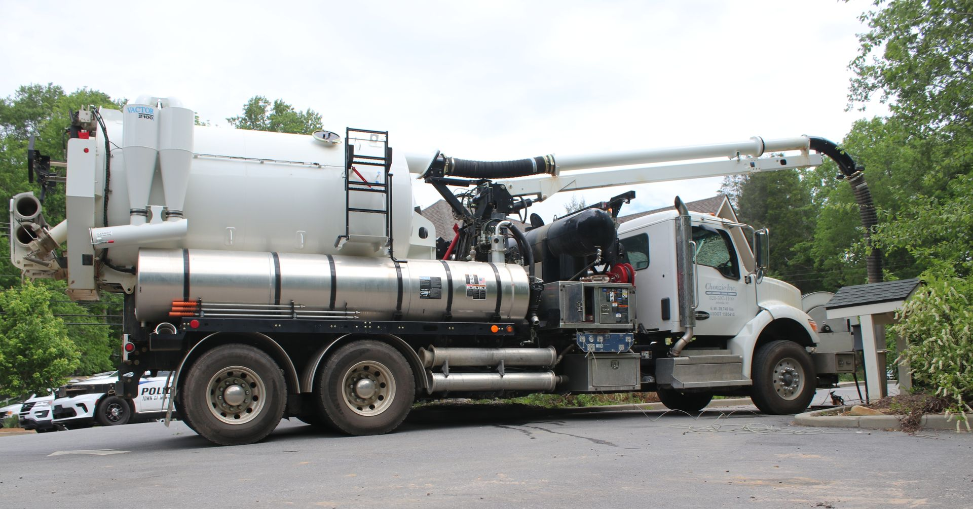 Large white industrial vacuum truck with hoses parked on a road among trees and cars.