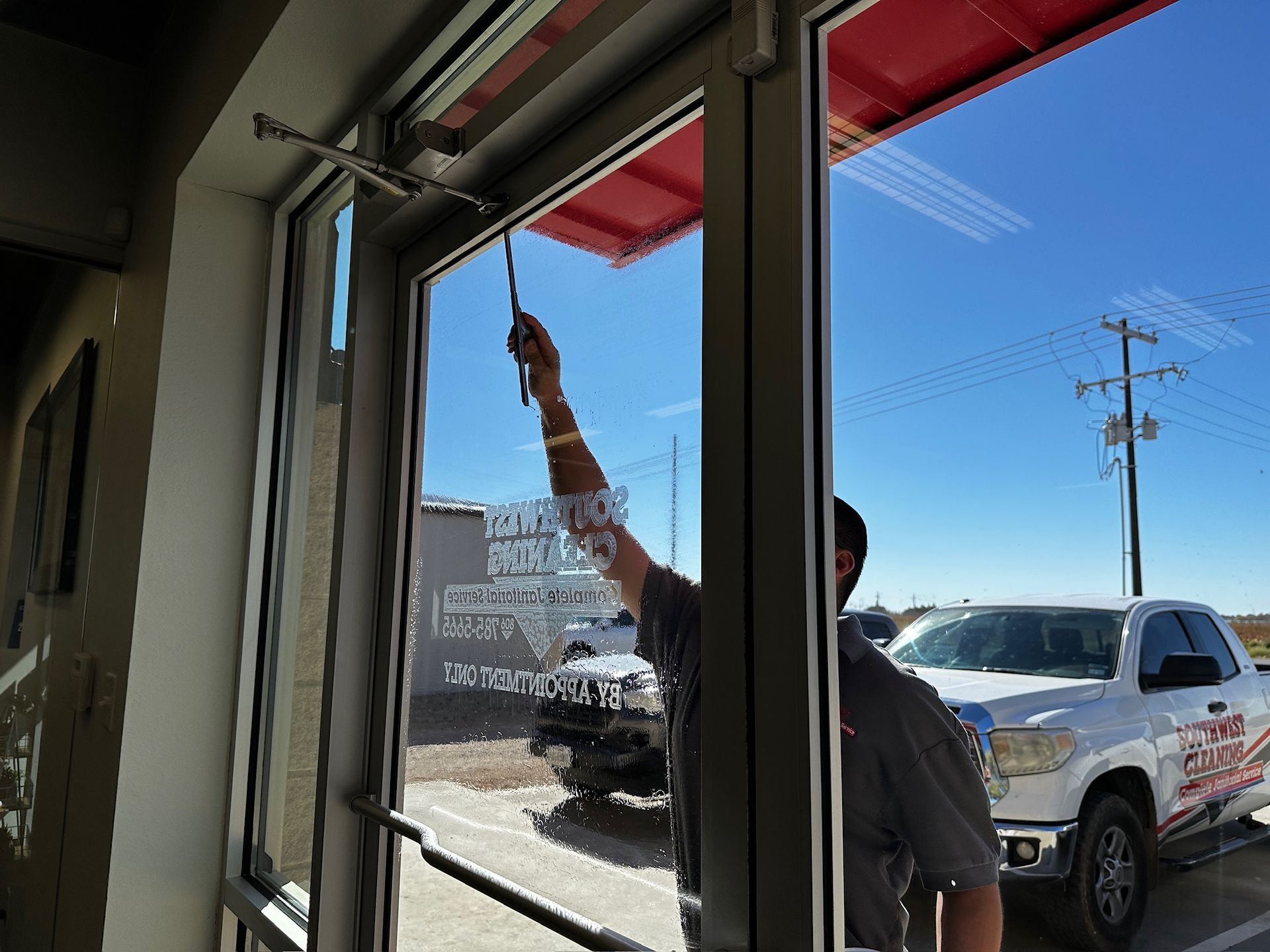 Person washing an interior window with a squeegee, with a bright street and pickup truck outside.