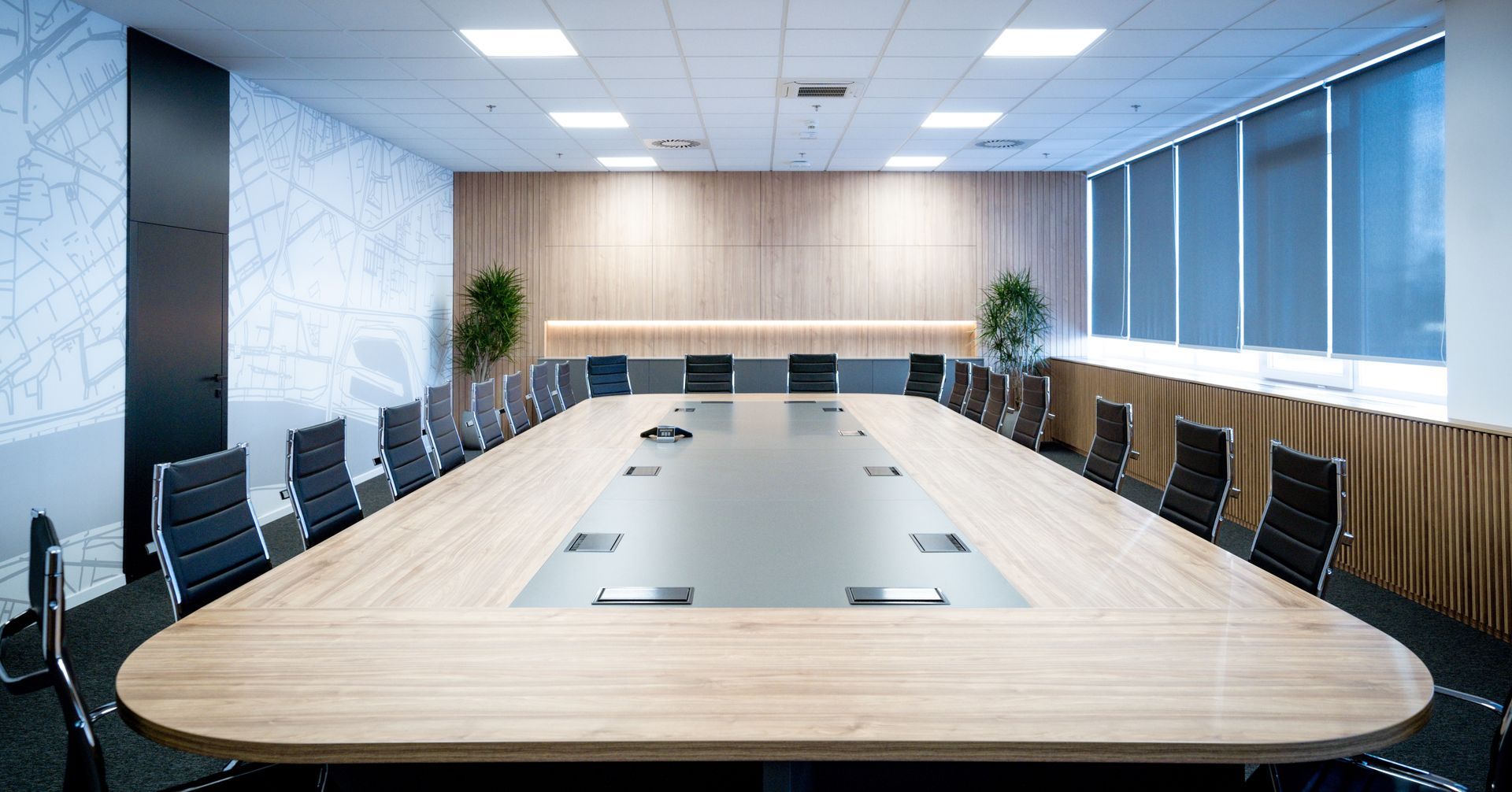 Modern conference room with a large oval table, black chairs, wood accent wall, and windows with blinds