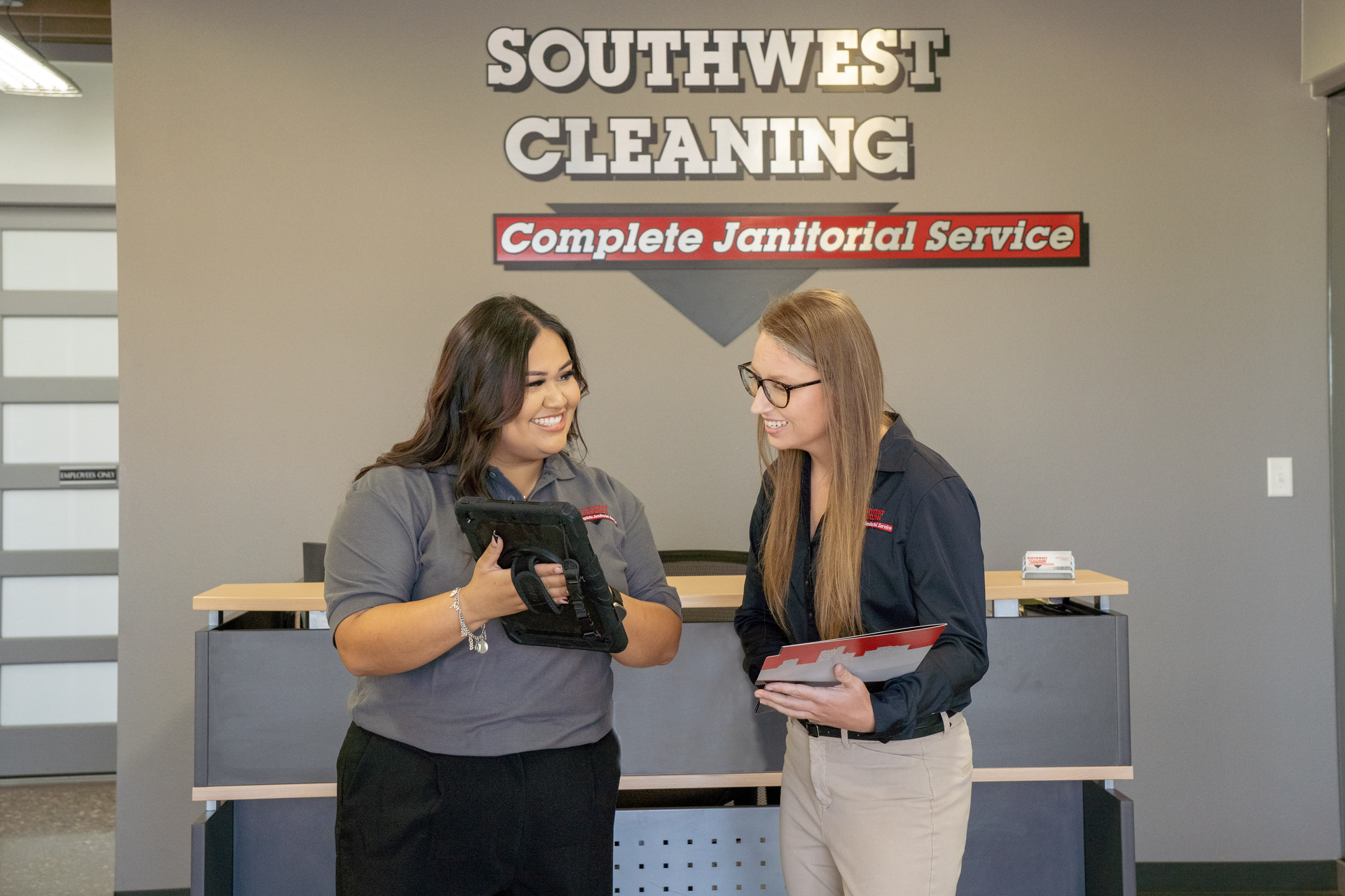 Two women in work uniforms, smiling, looking at tablets in an office setting with