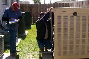 Two men inspecting a large beige cooling unit outdoors on a sunny day; one man is writing notes.