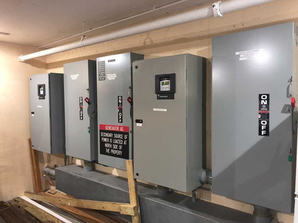 Electrical control panels mounted on a wooden wall, various sizes, gray, with labels and switches.