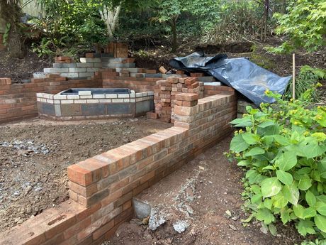 Brick wall construction outside in garden by brad with bushes and trees in background and some tools