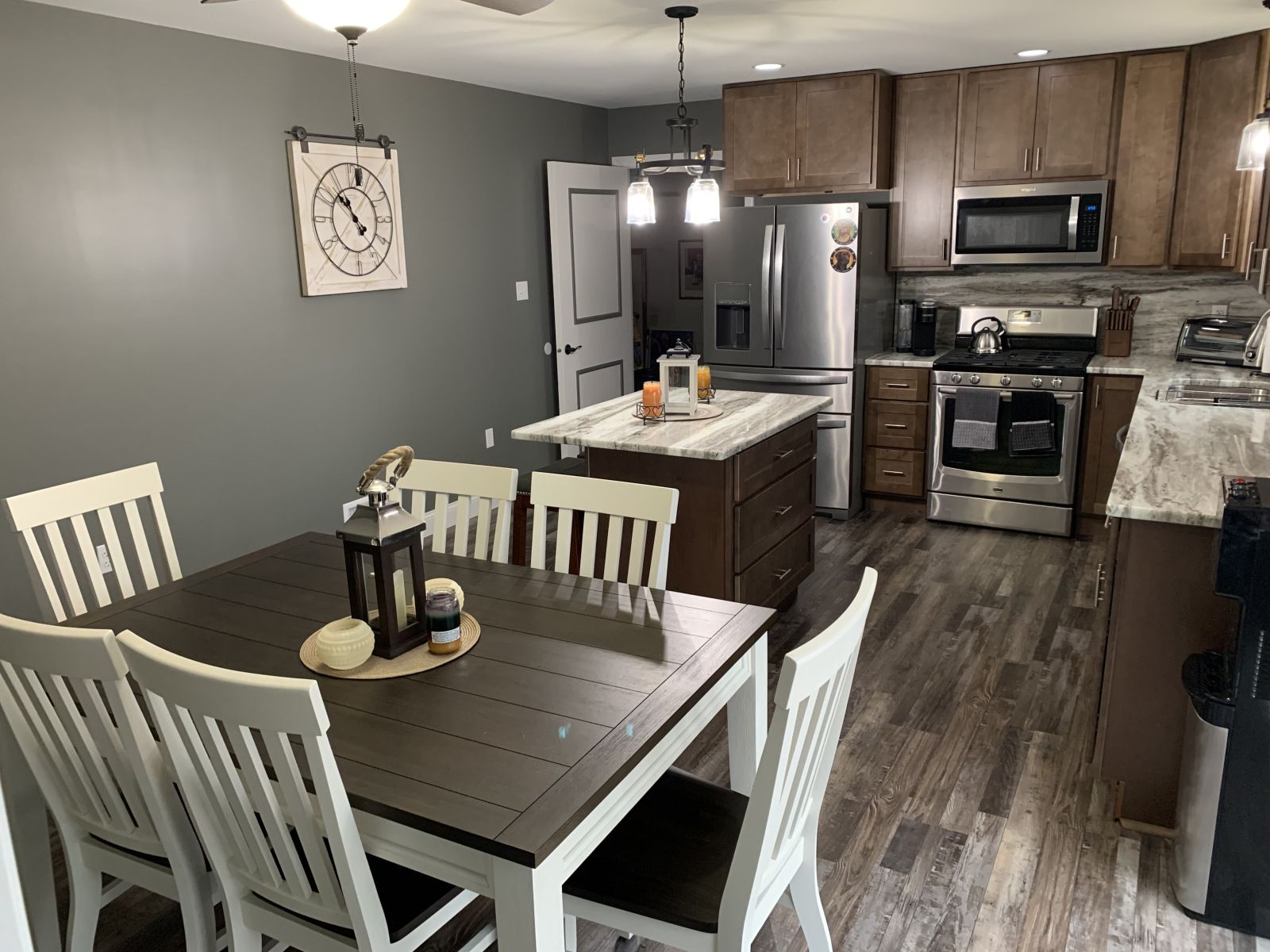 Kitchen with a dining table, island, and stainless steel appliances. Dark table, white chairs, gray walls, and brown cabinets.
