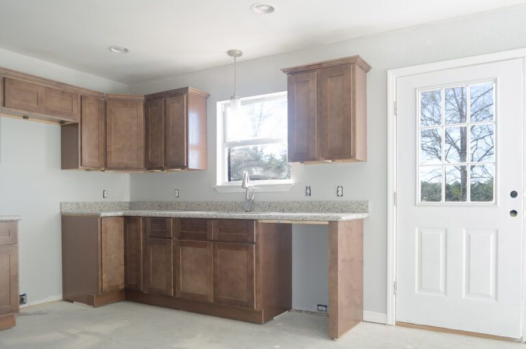 Kitchen with medium-toned wood cabinets, light countertops, a window, and a white door; unfinished flooring and walls.