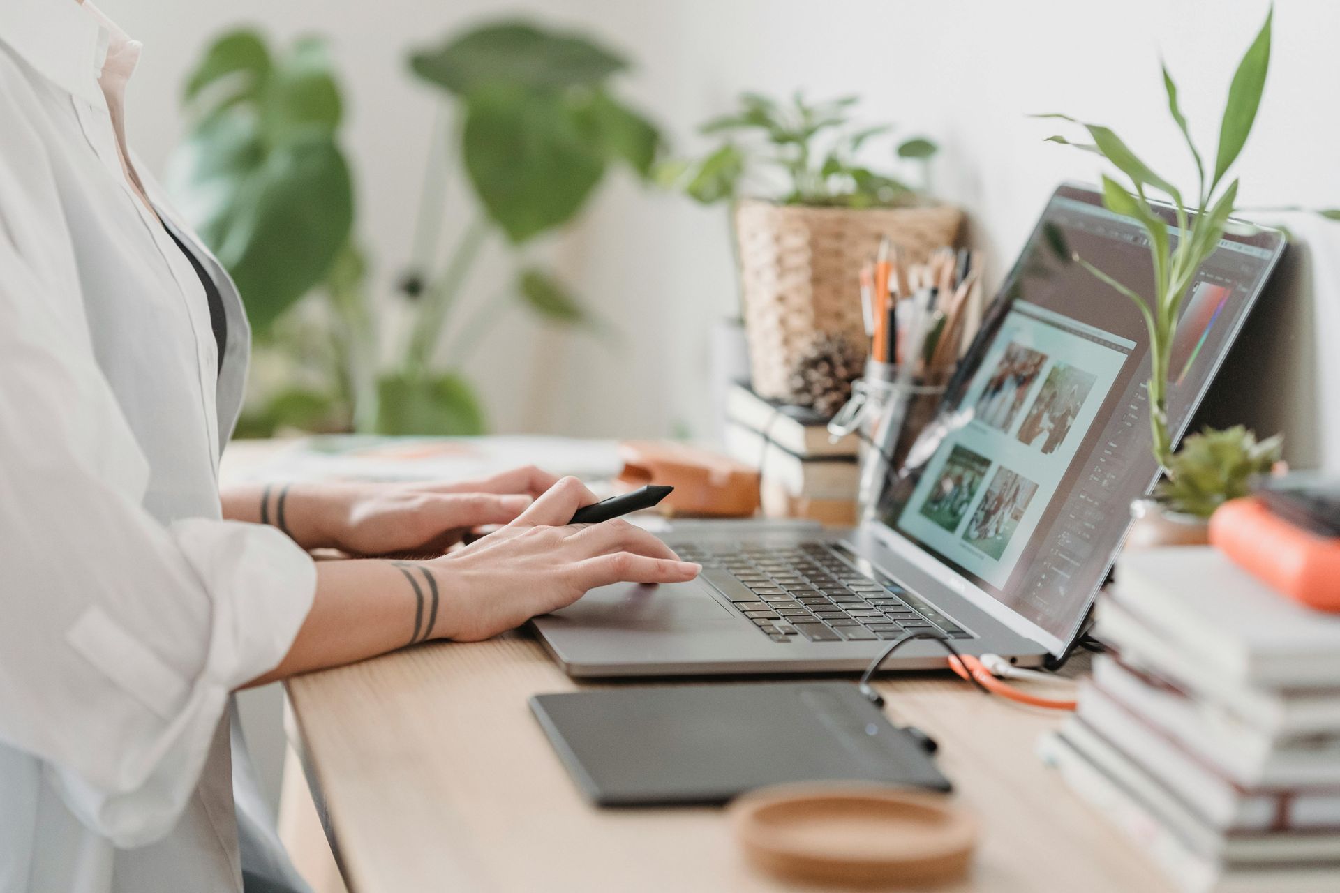 Woman working on laptop at desk, with plants and creative tools, indoor setting.