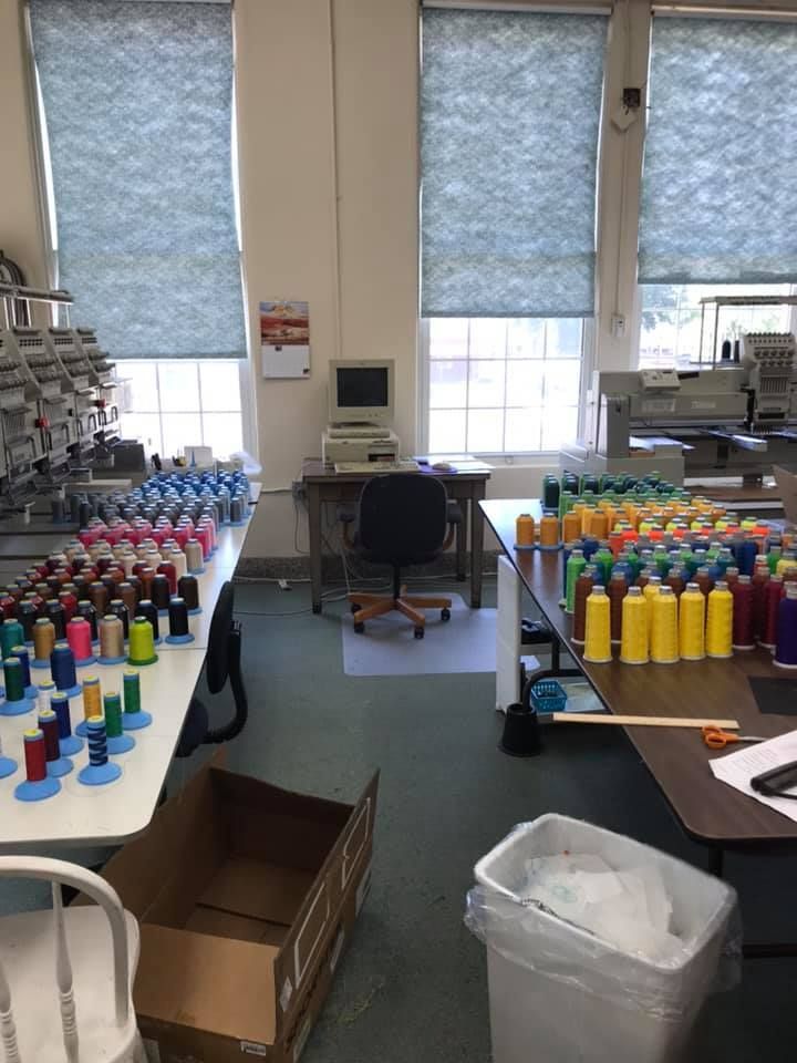 A sewing workspace with thread spools, computers, tables, and embroidery machines near large windows.