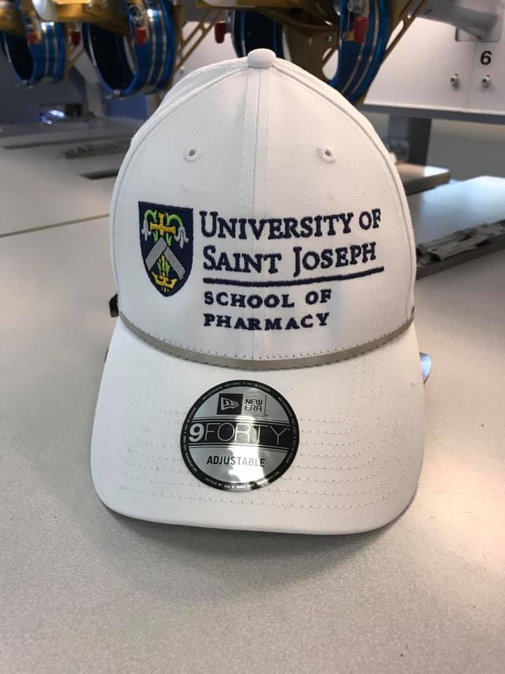 White New Era cap with University of Saint Joseph School of Pharmacy logo embroidered on the front.