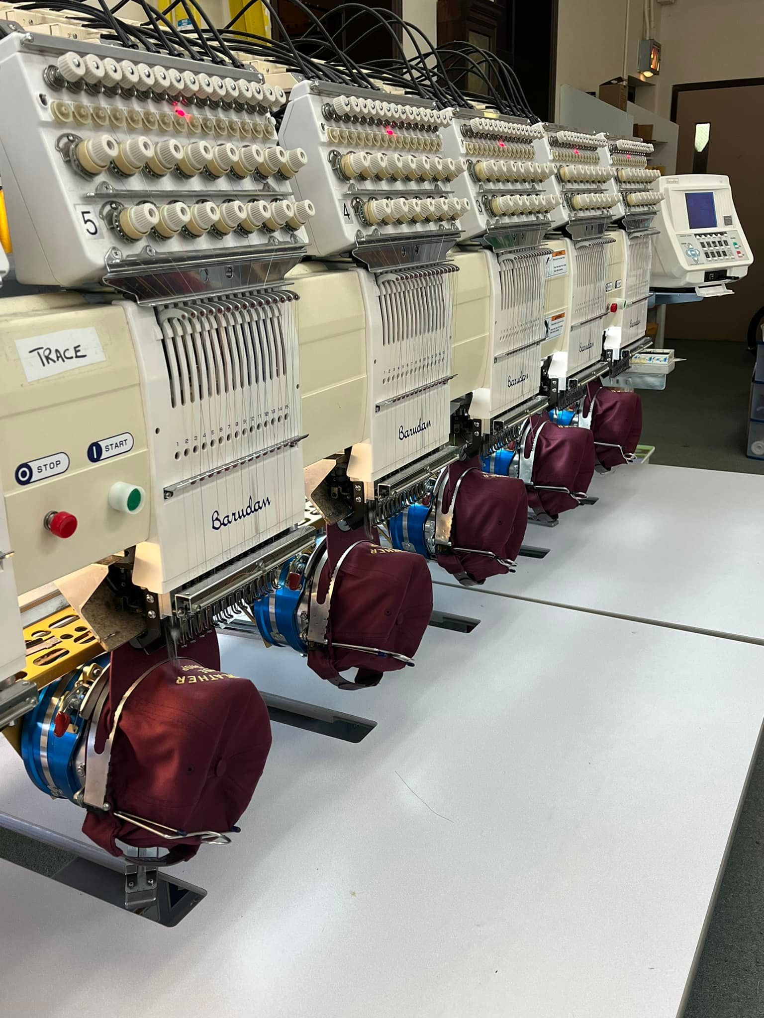 Row of industrial embroidery machines in a workshop, white and beige with numerous spools and colored thread covers.
