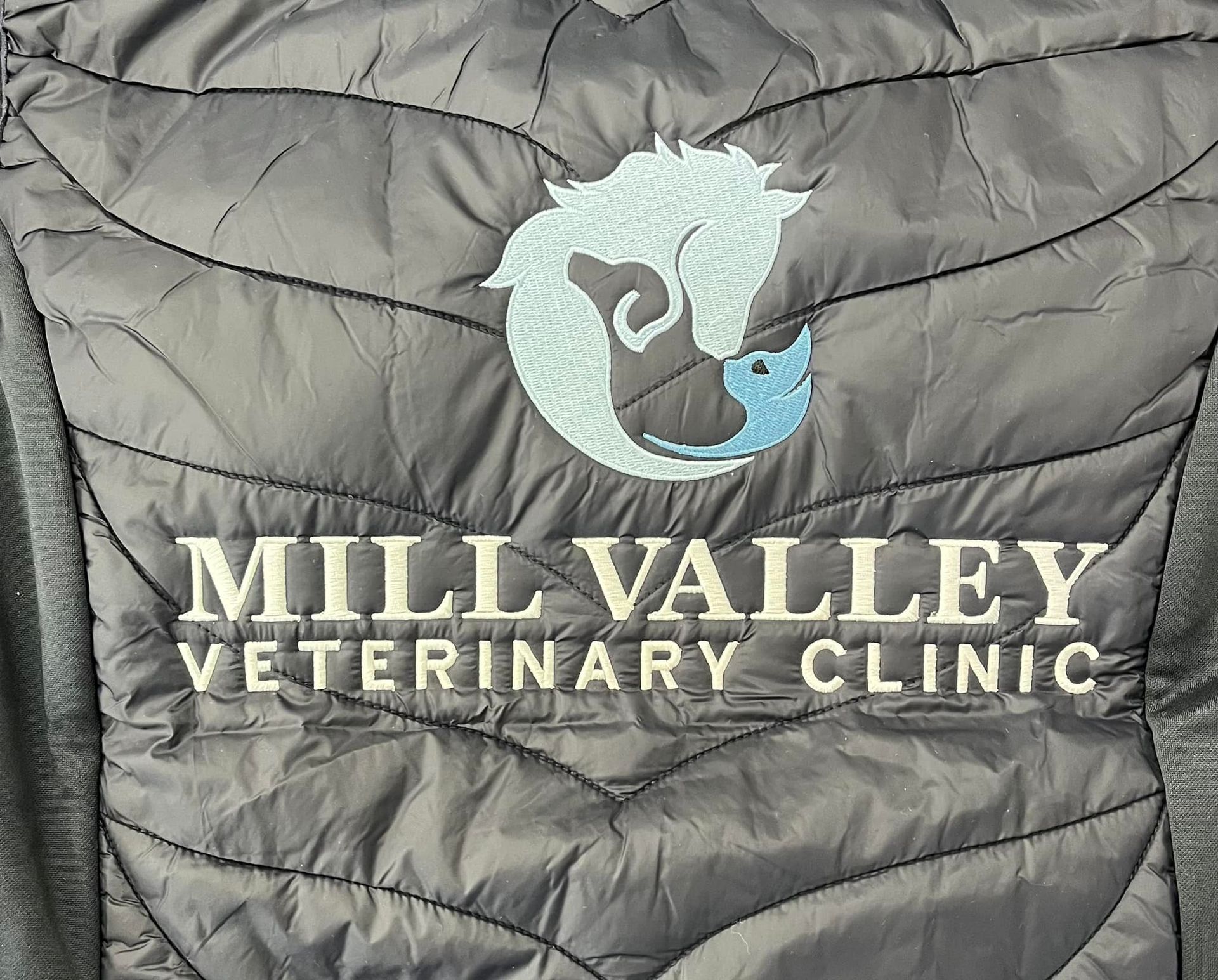 Dark gray jacket with light blue and white logo of horse head and text: Mill Valley Veterinary Clinic.