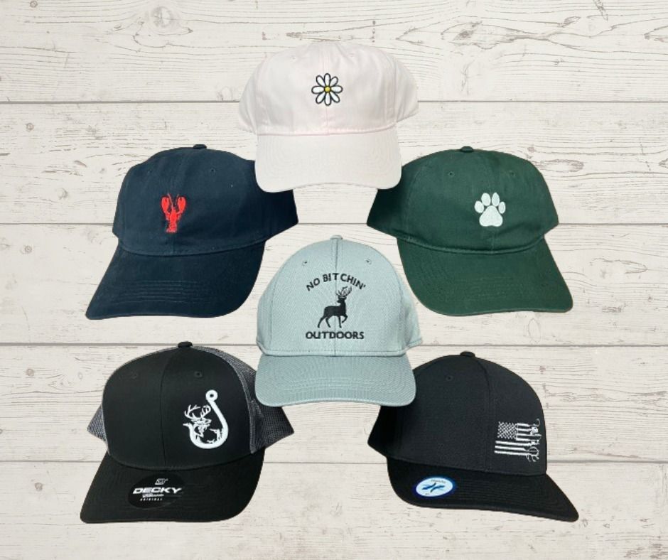 Seven baseball caps of different colors, with embroidered designs, arranged in a semi-circle on a wood-grain background.