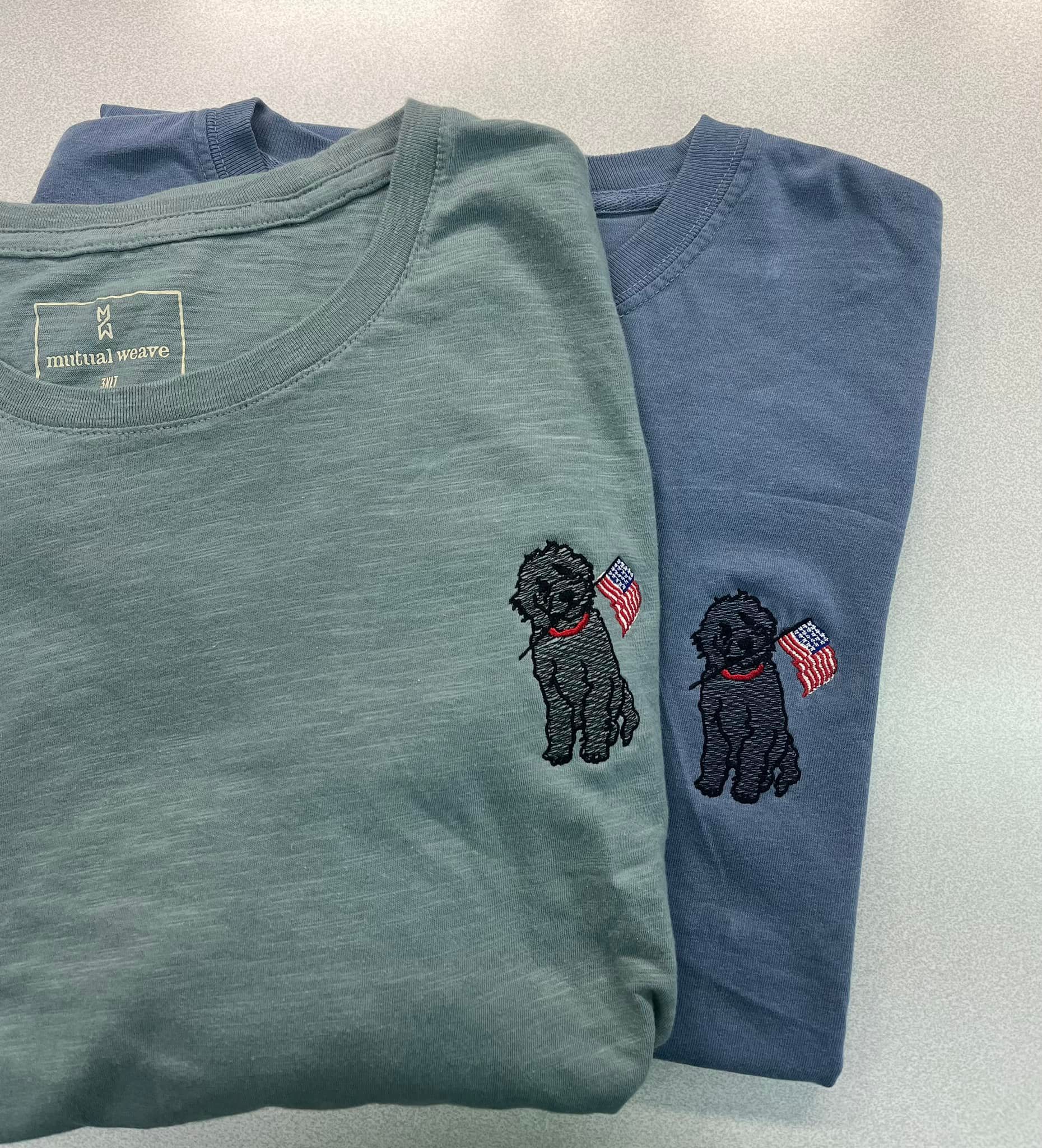 Two folded t-shirts, green and blue, with embroidered black dog holding an American flag.
