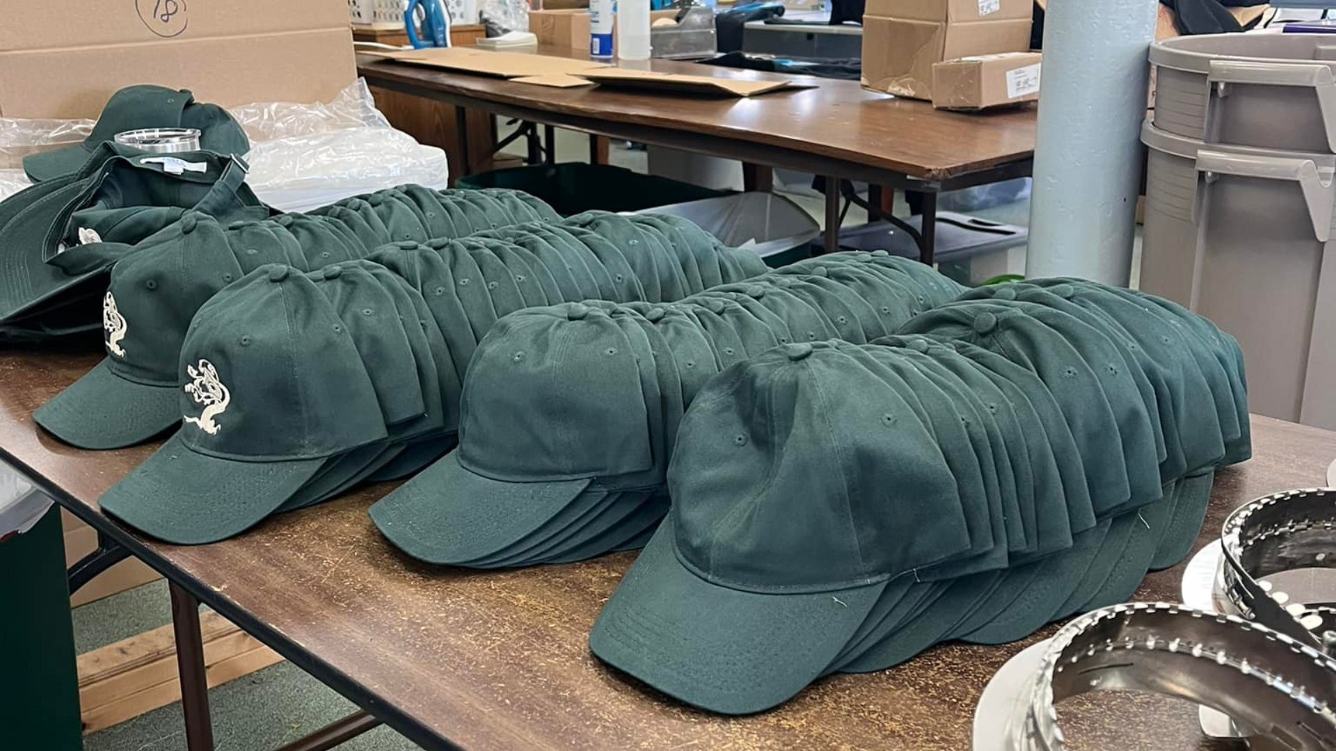 Green baseball caps with white logos stacked on a brown table.