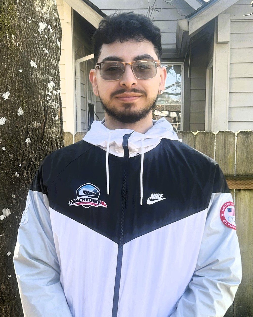 A man wearing sunglasses and a nike jacket is standing in front of a house.