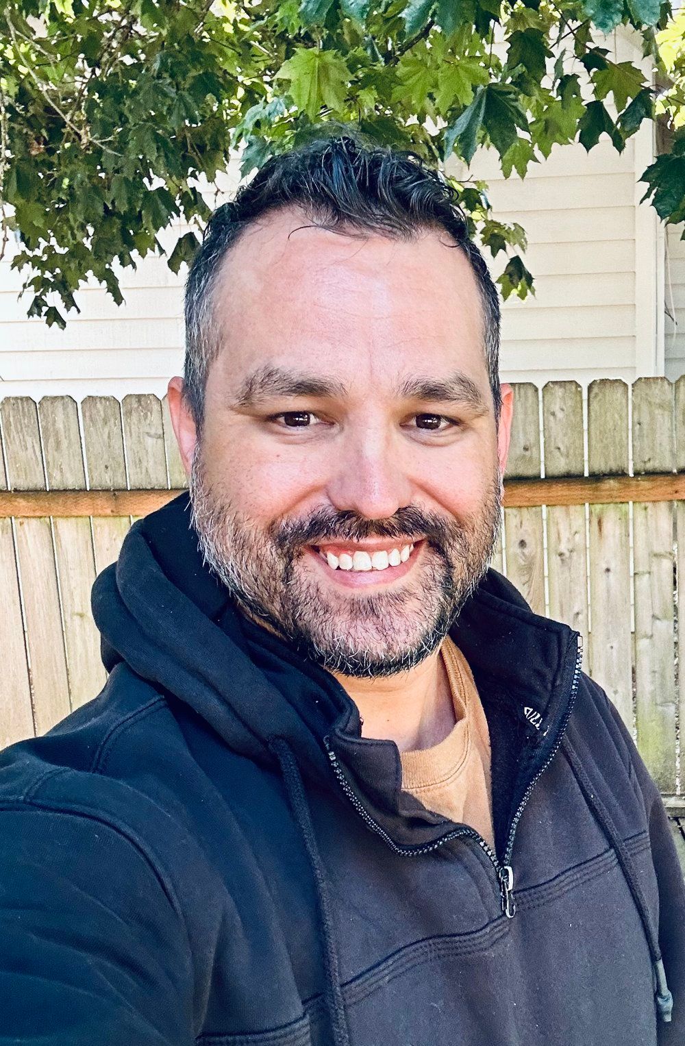 A man with a beard is smiling for a selfie in front of a wooden fence.