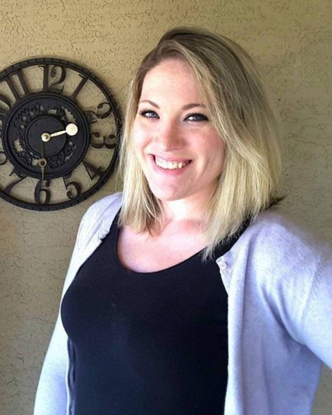 A woman is smiling in front of a large clock on the wall.