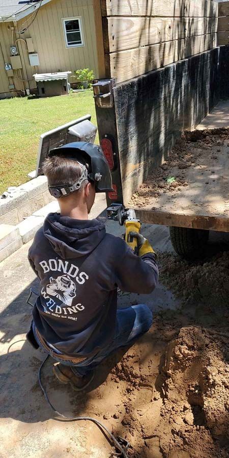 A man wearing a welding helmet is kneeling down in the dirt next to a truck.