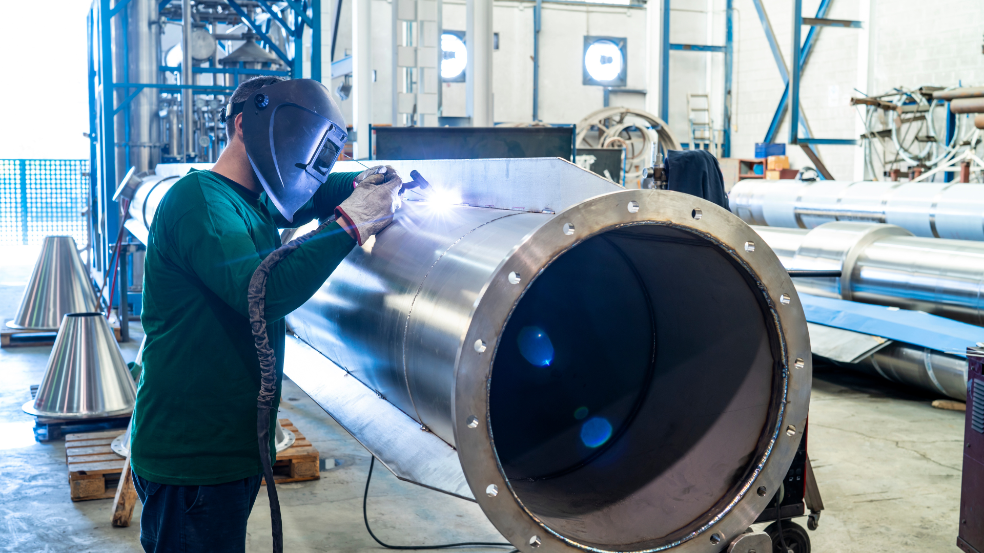 A man is welding a large metal pipe in a factory.