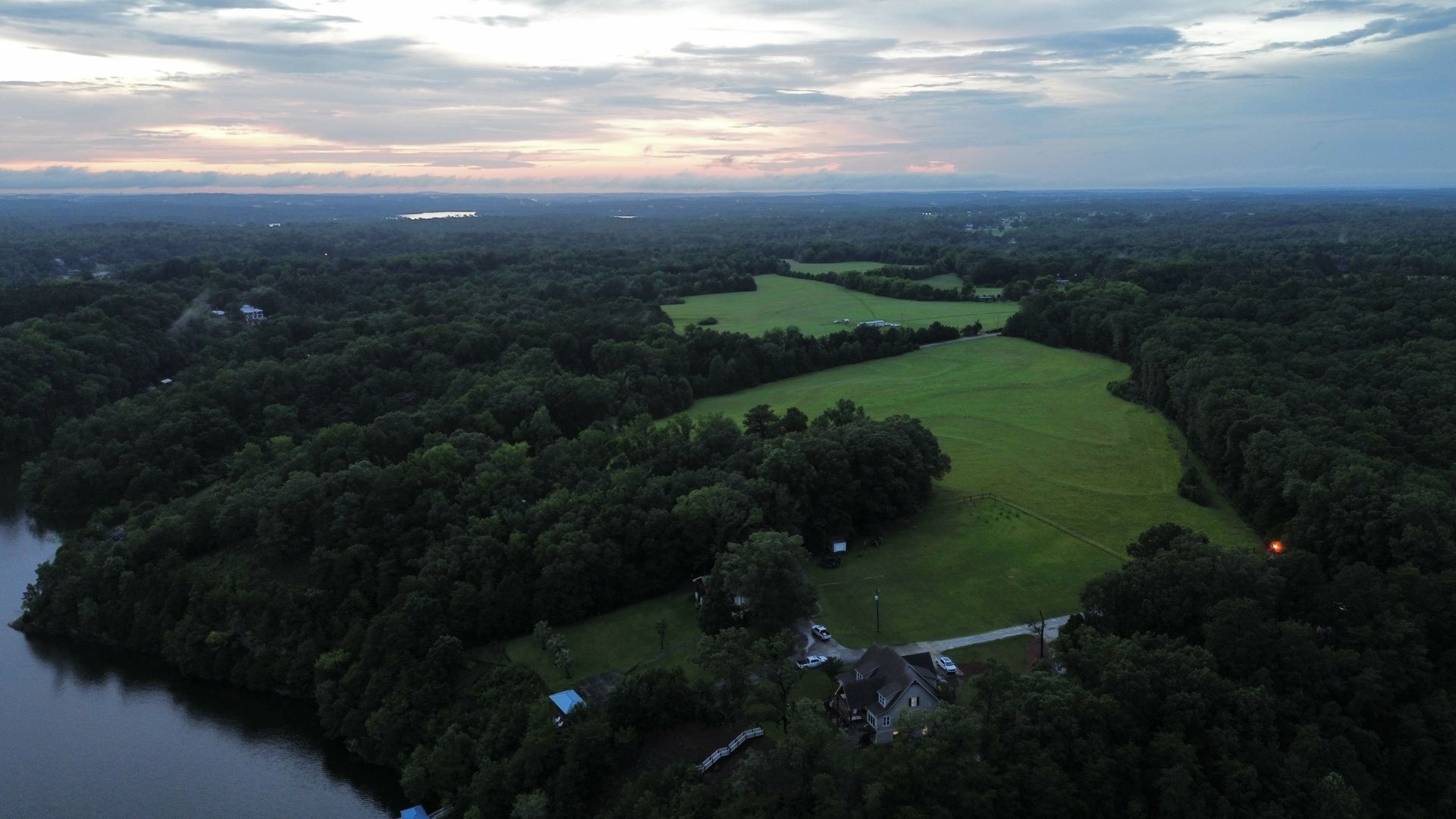 Aerial view of a grassy field surrounded by dark green trees under a cloudy sky.